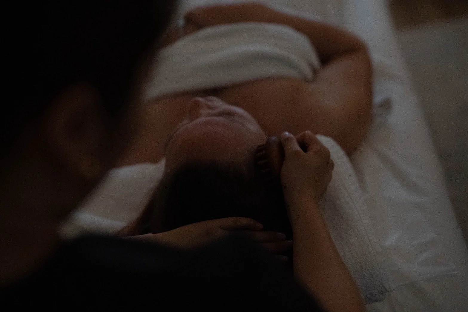 A person lying on a bed with one hand resting on their face and the other on their neck, wearing a white top, with a blanket covering their lower body.