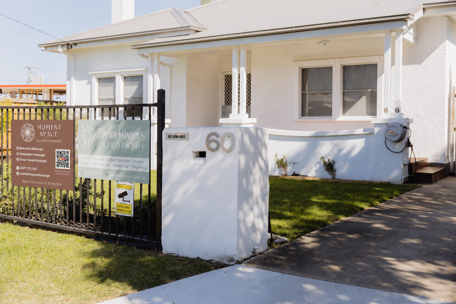 White house with a small front yard and black metal fence, displaying signs for wellness services, located at 60 on the street. Signs include 'Moment Space Wellness Massage' and 'Twin Rivers Osteopathy'.