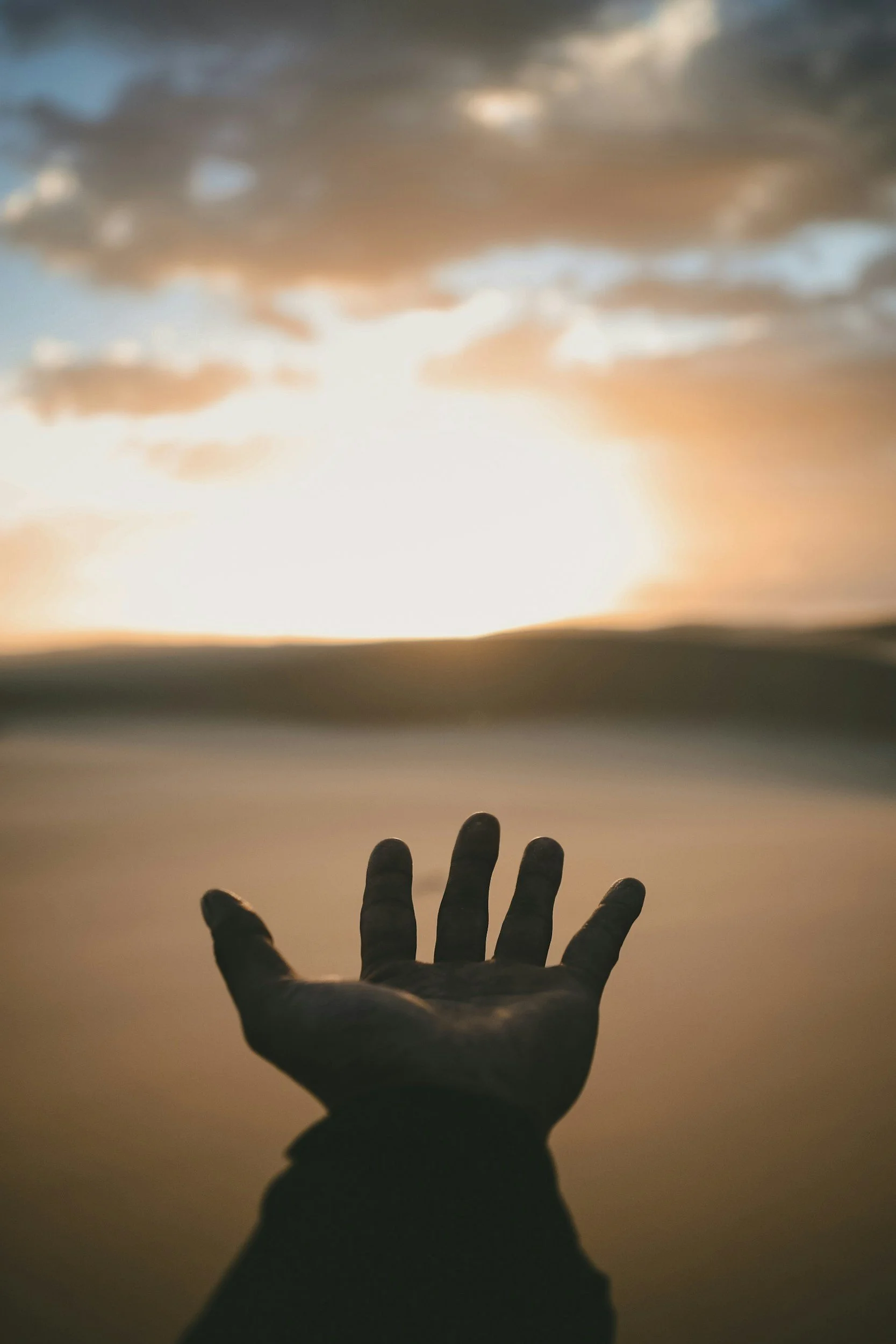 A hand reaching out toward the sky with clouds and a setting sun in the background.