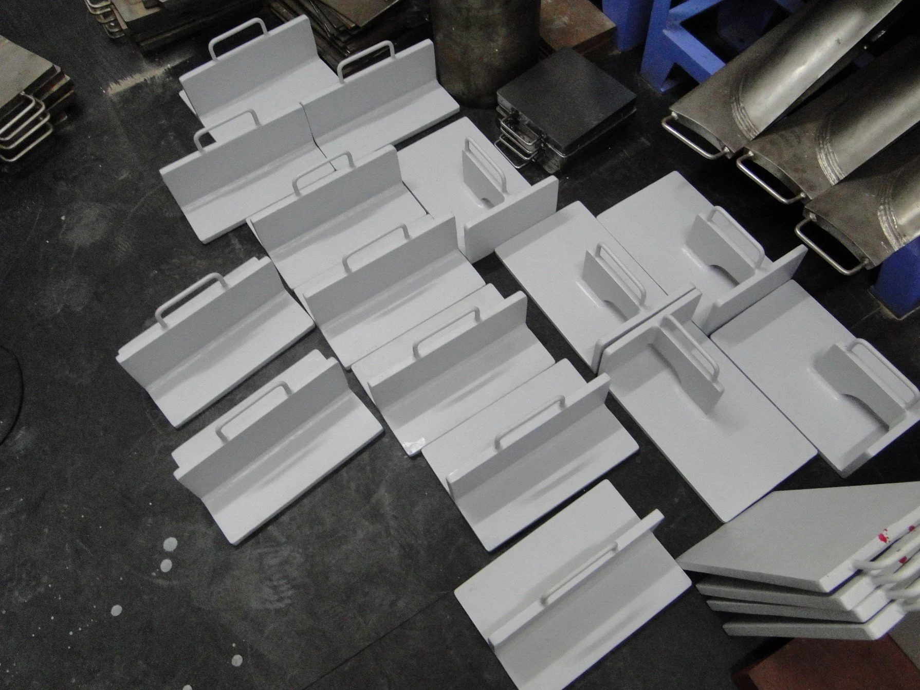 A collection of white metal or plastic trays with handles, arranged on a dark floor or table. Some objects are stacked and others are laid out flat. There are also black objects and metallic pipes or metal tubes nearby.