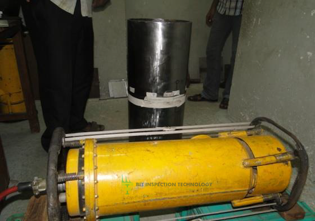 A hydraulic or pneumatic cylinder with a yellow body and black end caps on a table, with a large black drum and a person standing nearby.