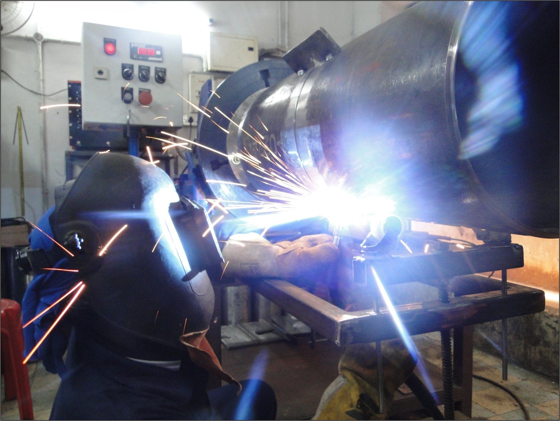 A person welding metal pipes in a workshop, with sparks flying and welding equipment visible.