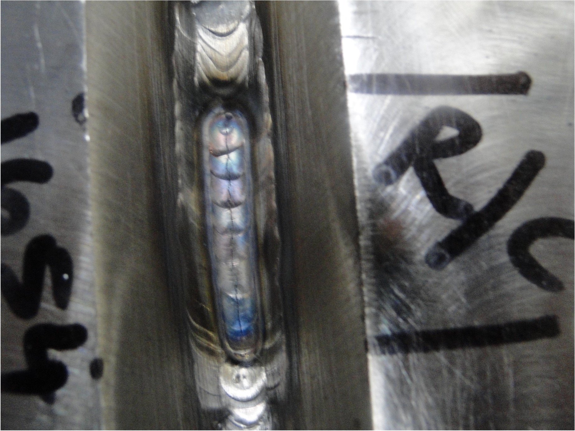 Close-up of a metal surface with handwritten black marker writing, showing a weld bead or seam with a slight rainbow discoloration.