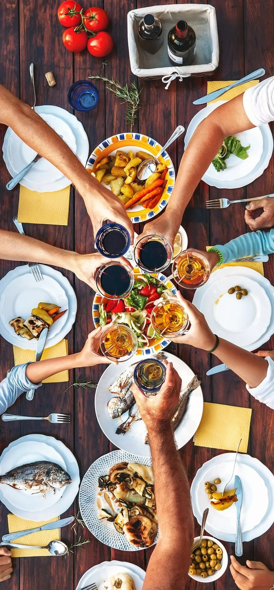 A top-down view of a dinner party with various dishes including fish, salad, roasted vegetables, and tomatoes, along with glasses of red wine being raised for a toast on a wooden table.