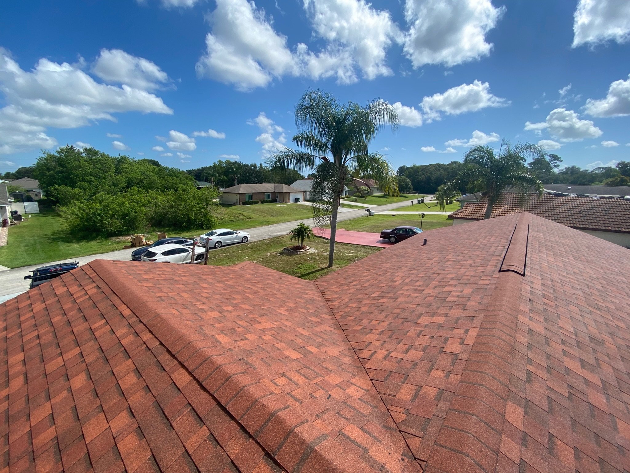 A suburban neighborhood with houses, trees, parked cars, and a blue sky with scattered clouds.