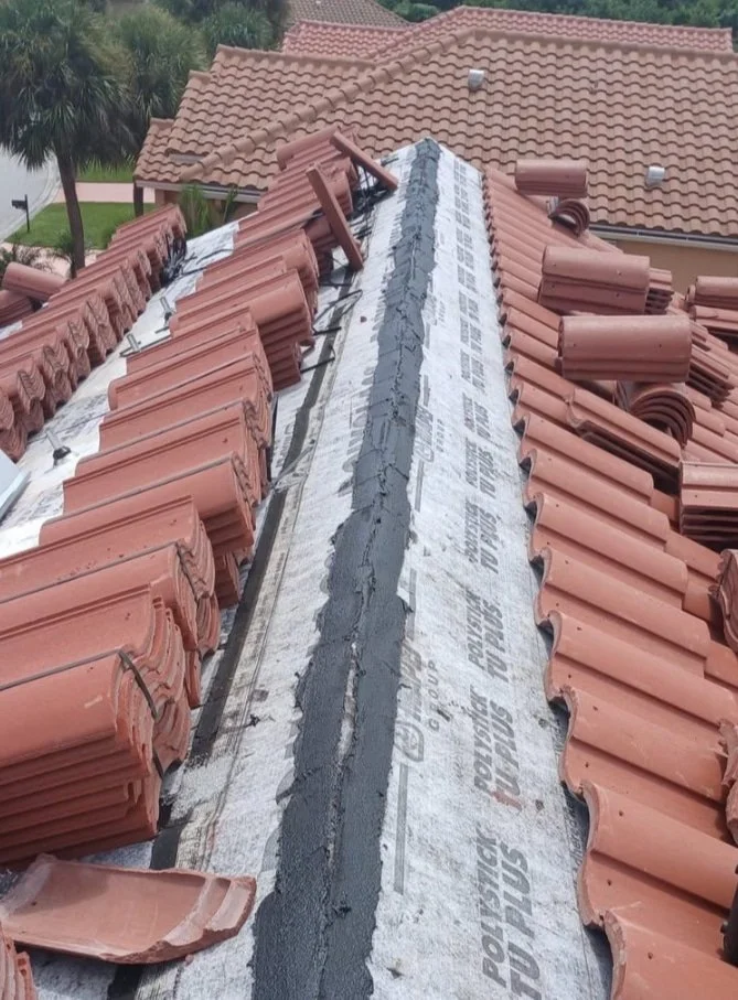 Stacks of red roof tiles and roofing paper line both sides of a roof ridge during construction, with sealant in the center.
