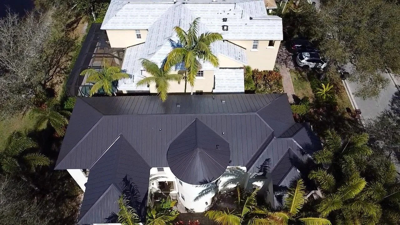 An aerial view of a large house with a black metal roof surrounded by palm trees and greenery.