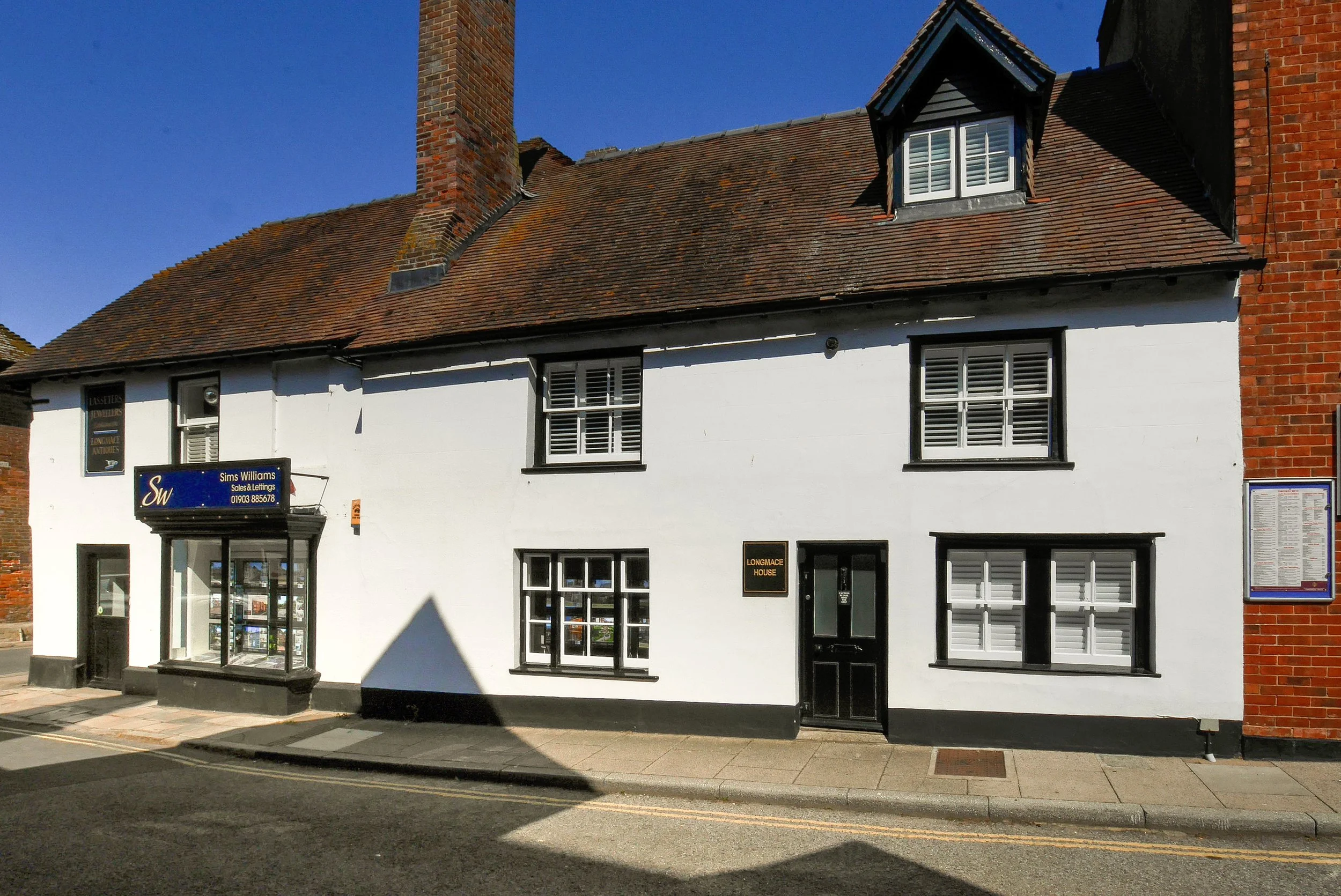 White building with black window frames and a sign that reads 'Sims Williams Sales & Lettings' in blue, located on a street with a sidewalk and road. The roof is made of reddish-brown tiles, and there are brick accents on the building, with a clear blue sky in the background.