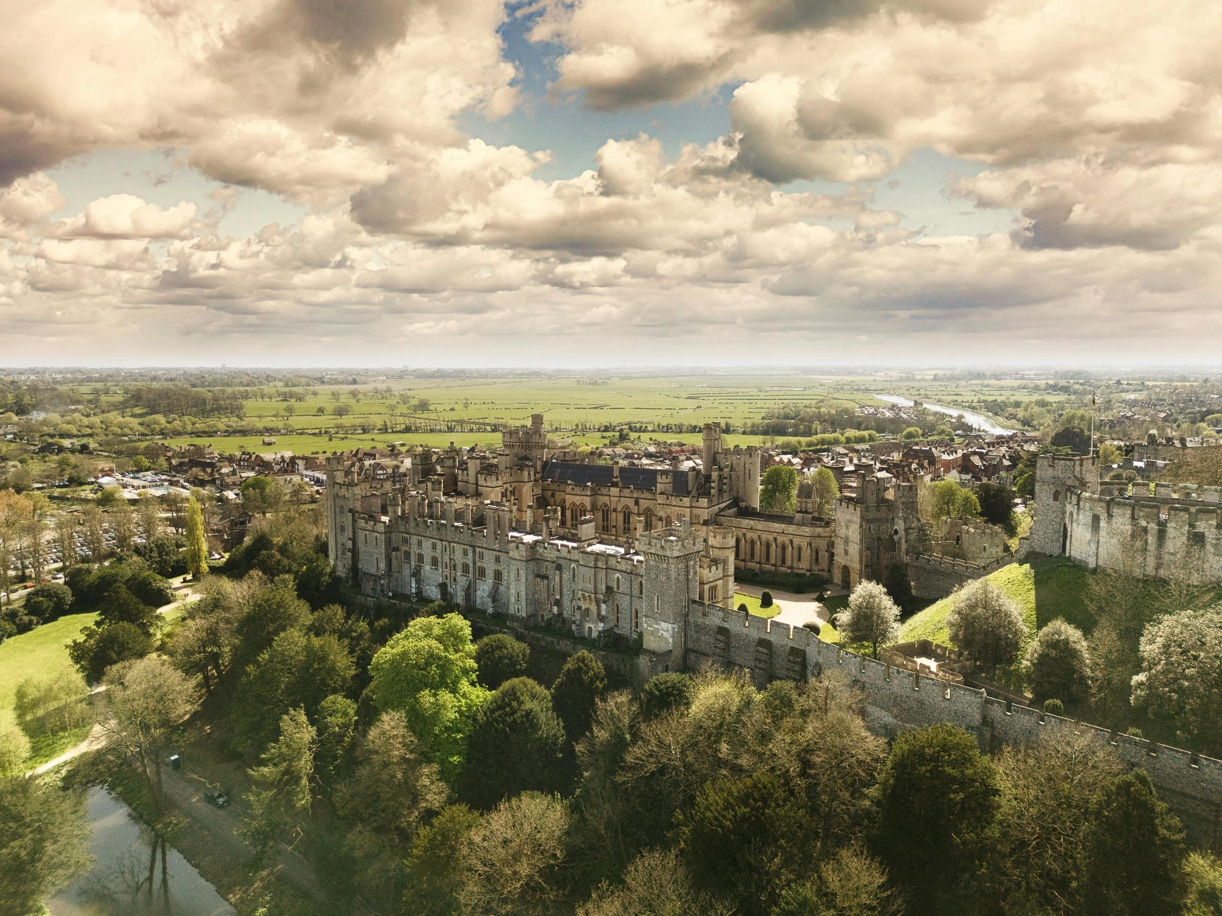 A panoramic view of a castle surrounded by green trees, with a town and river in the distance under a cloudy sky.