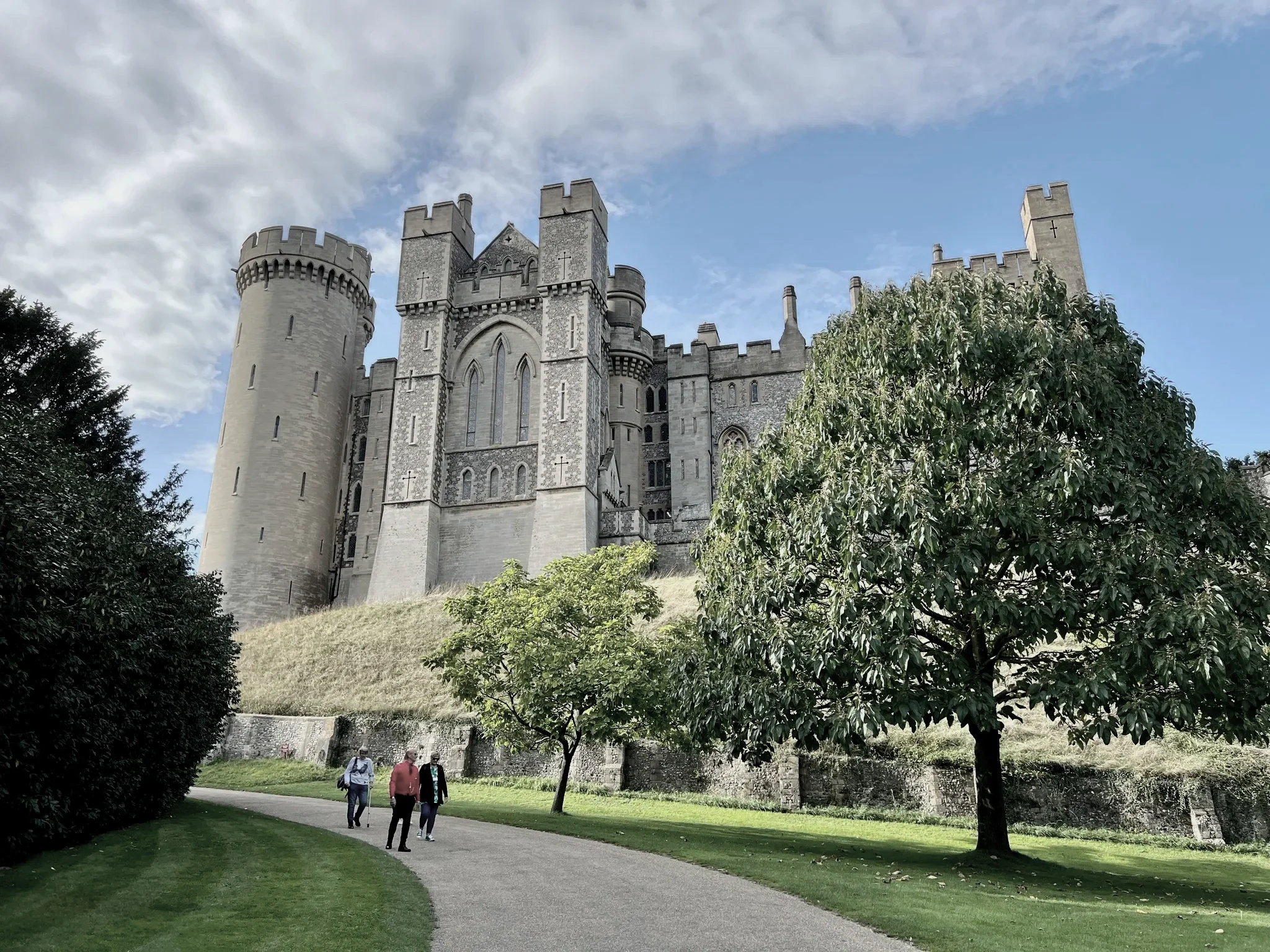 Four people are walking along a paved path in a park with a castle in the background. The castle has tall towers and stone walls, with trees and green grass surrounding it.