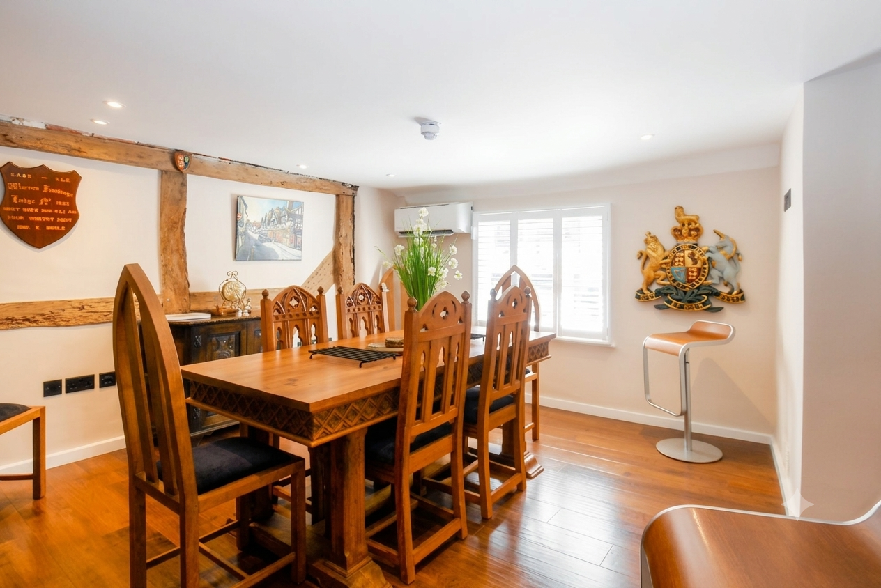 Dining room with a wooden dining table, tall chairs, a black sideboard, a large plant centerpiece, a coat of arms on the wall, and a window.