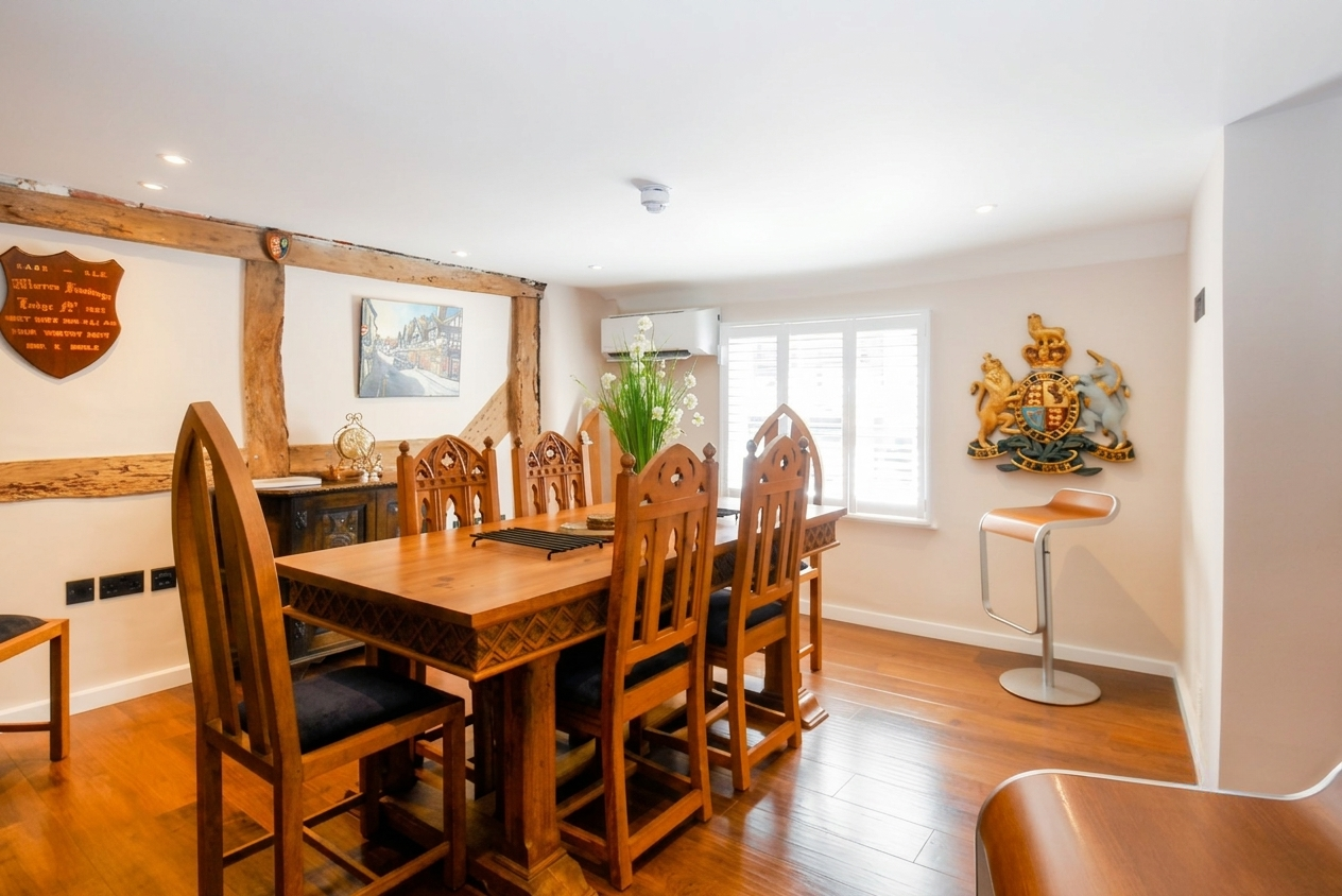 Dining room with a wooden table, six high-backed chairs, a large flower arrangement, a coat of arms crest on the wall, a painting of city street, and windows with blinds.
