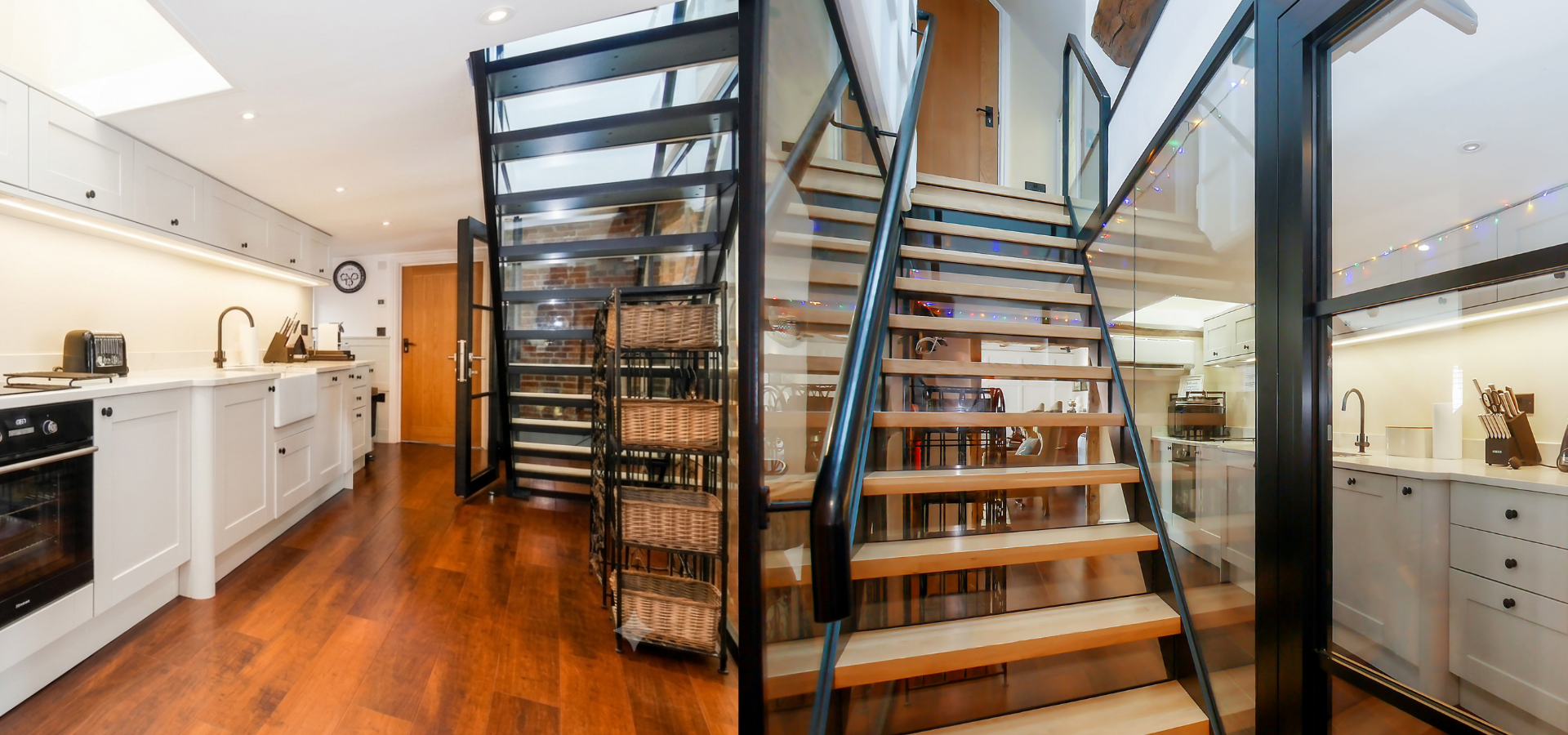 Interior view of a modern kitchen with white cabinets, a wooden floor, a staircase with wooden steps and black metal railing, and a glass wall separating the kitchen from the staircase area. The kitchen features appliances and utensils, with some decorative string lights.