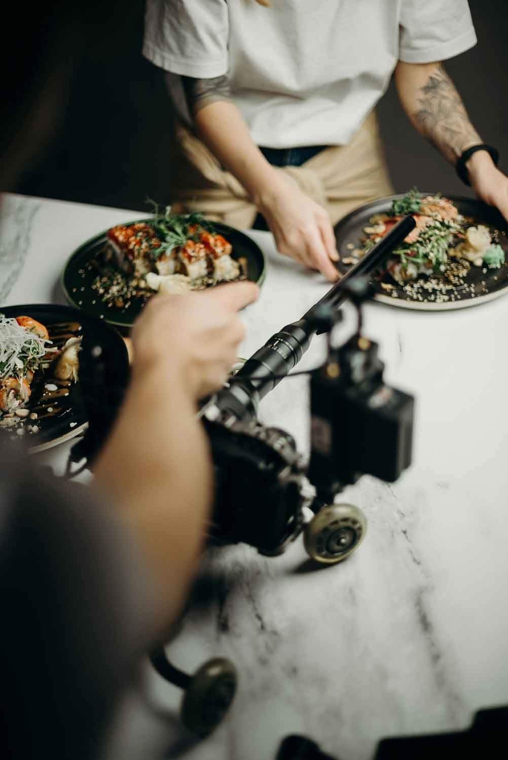A person shooting a food photo of sushi plates on a white table using a camera on a tripod.