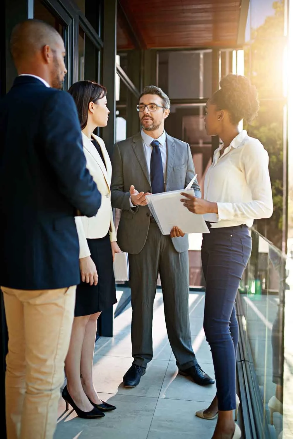 Four business professionals having a discussion in an office lobby with glass walls and a scenic outdoor view.