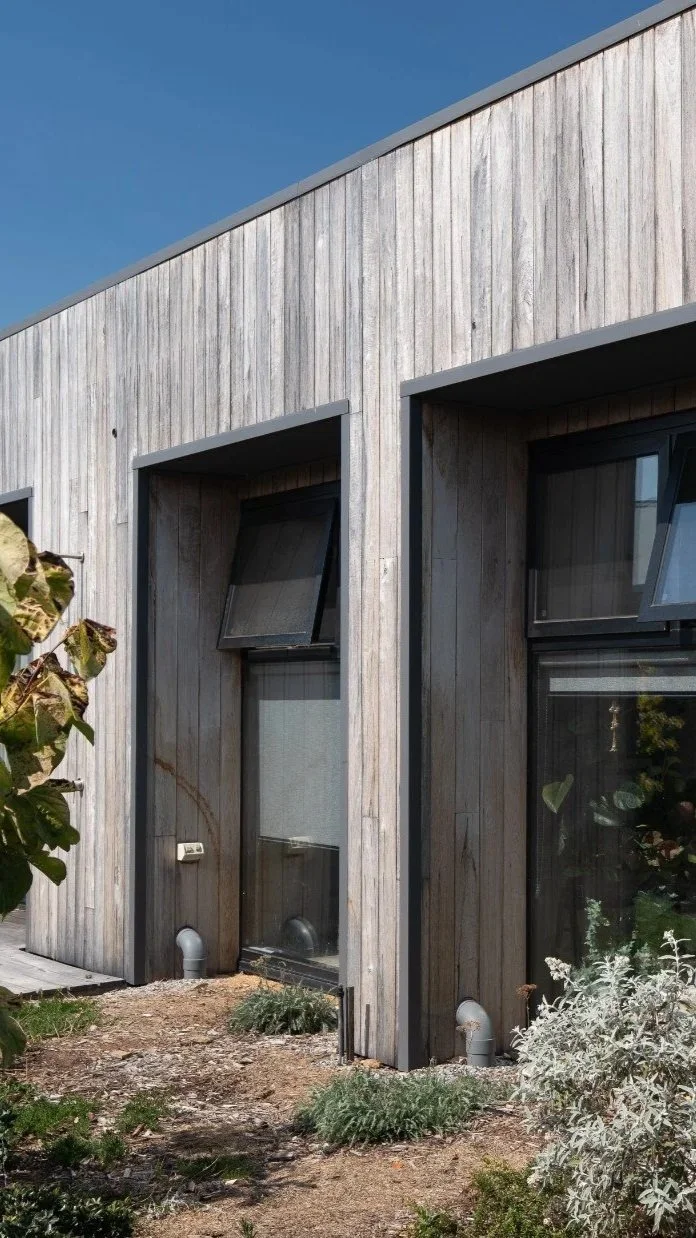 Modern house with wood siding, multiple large windows, and a garden with plants and shrubs.