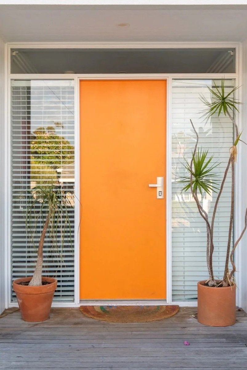 Front porch with an orange door, two potted plants on either side, and a doormat on a wooden deck.