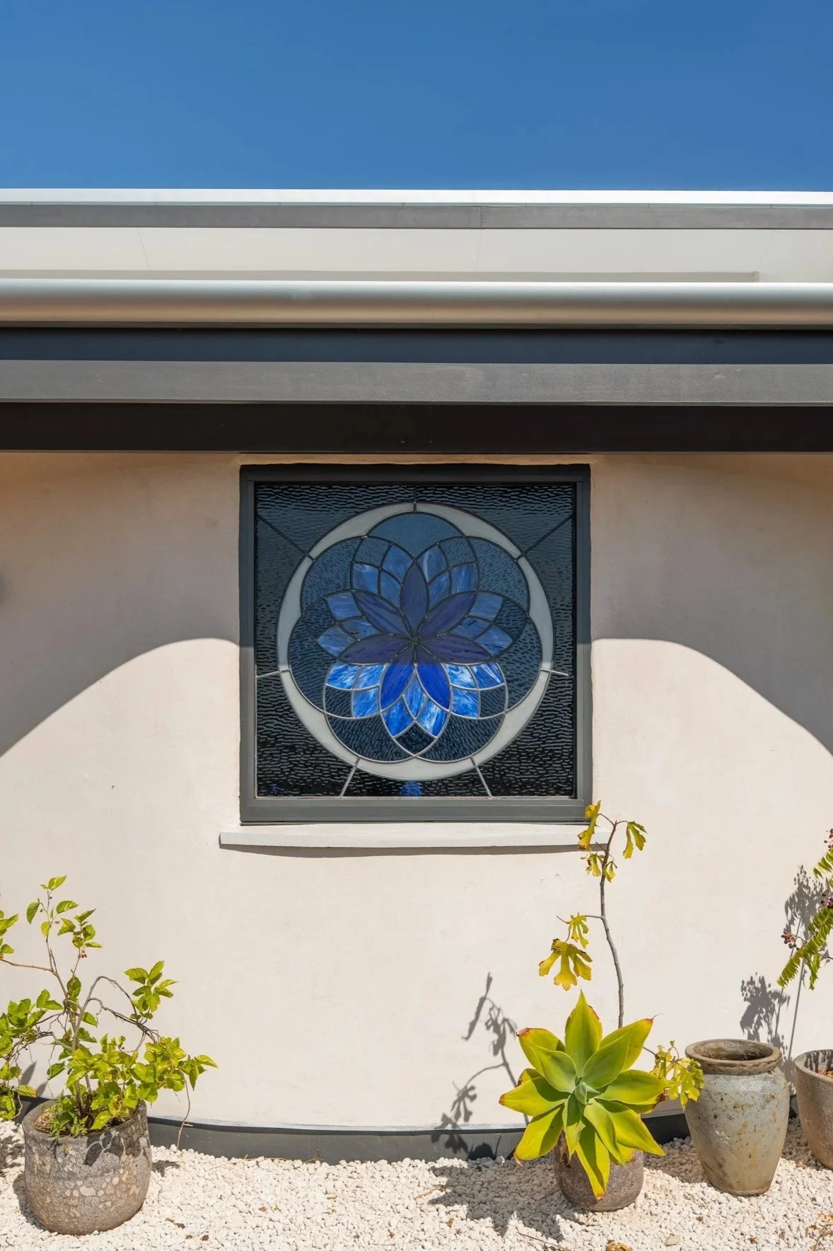 A white building with a stained glass window featuring a blue flower design, surrounded by potted plants and a gravel garden ground under a clear blue sky.