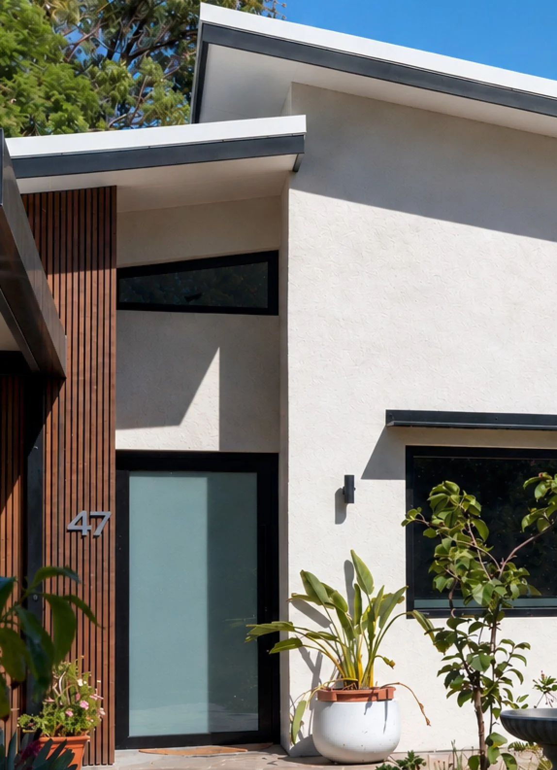 Modern house exterior with white walls, black window frames, and wooden accent wall, featuring house number 47, surrounded by potted plants and greenery under a clear blue sky.