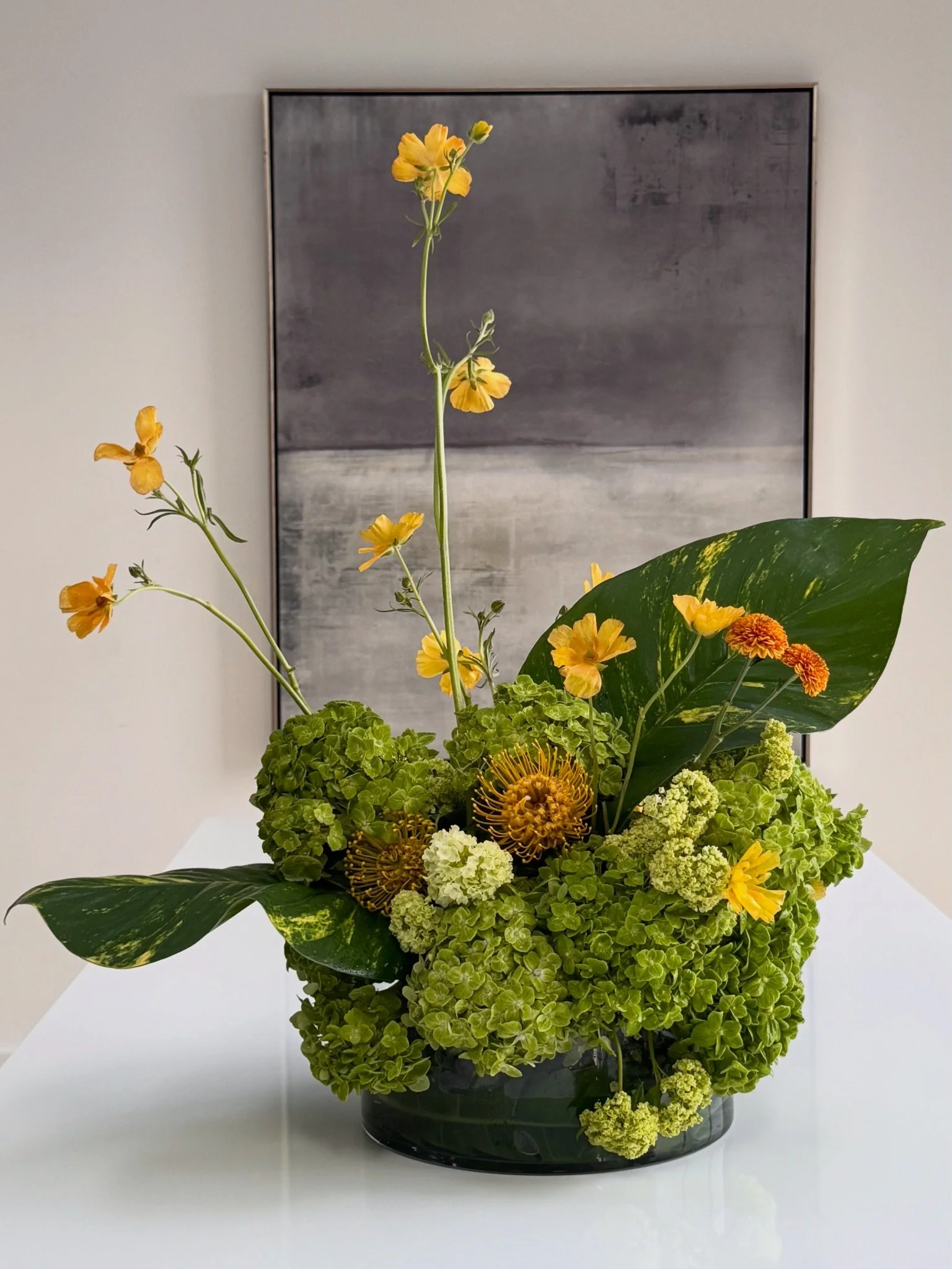 A floral arrangement with yellow, orange, and green flowers and leaves in a black container, standing on a white surface against a white wall with a black and white framed abstract art piece.