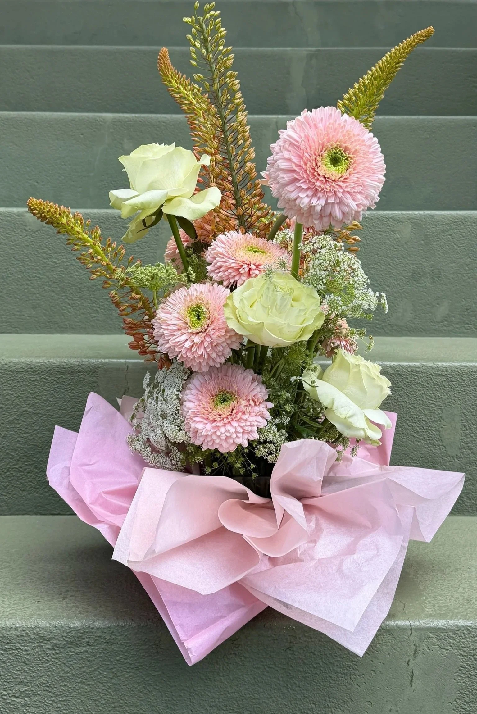 A bouquet of pink and white flowers, including pink gerbera daisies, white roses, and baby's breath, wrapped in pink paper and set against a green textured background.
