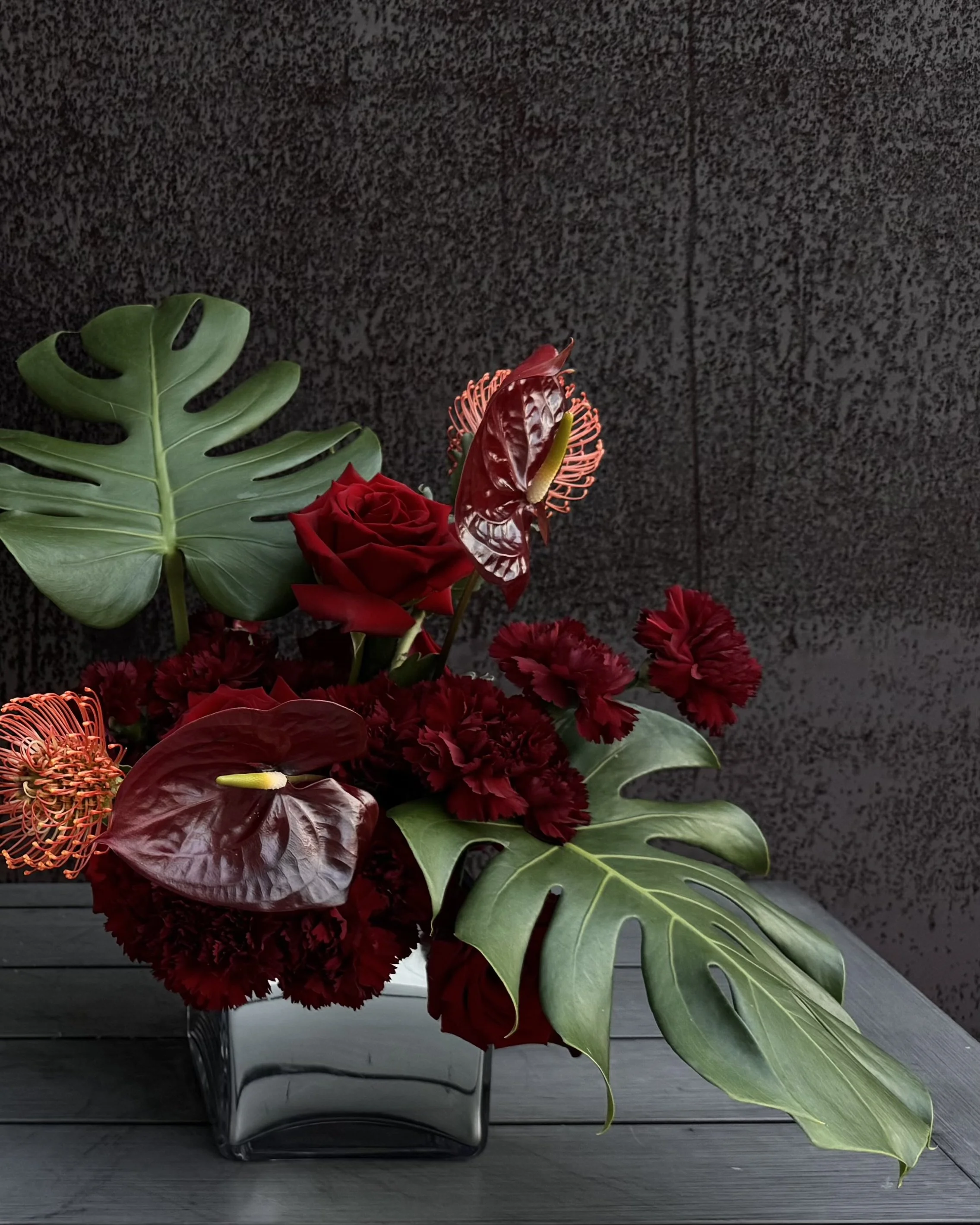 A floral arrangement in a black container with green monstera leaves, red roses, red anthuriums, and red carnations against a dark textured background.
