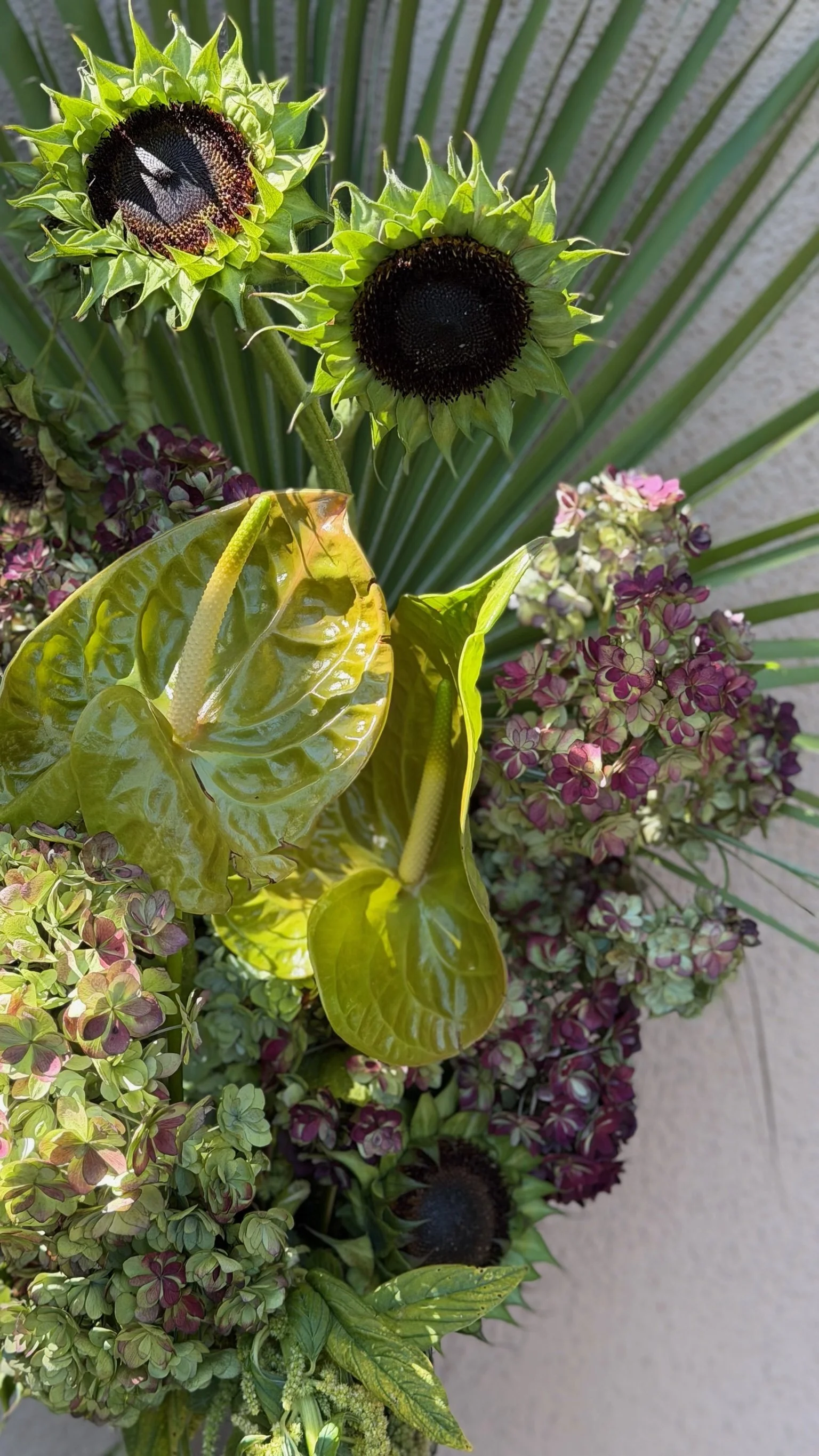 Close-up of a arrangement of sunflowers, green anthuriums, purple hydrangeas, and large green leaves.