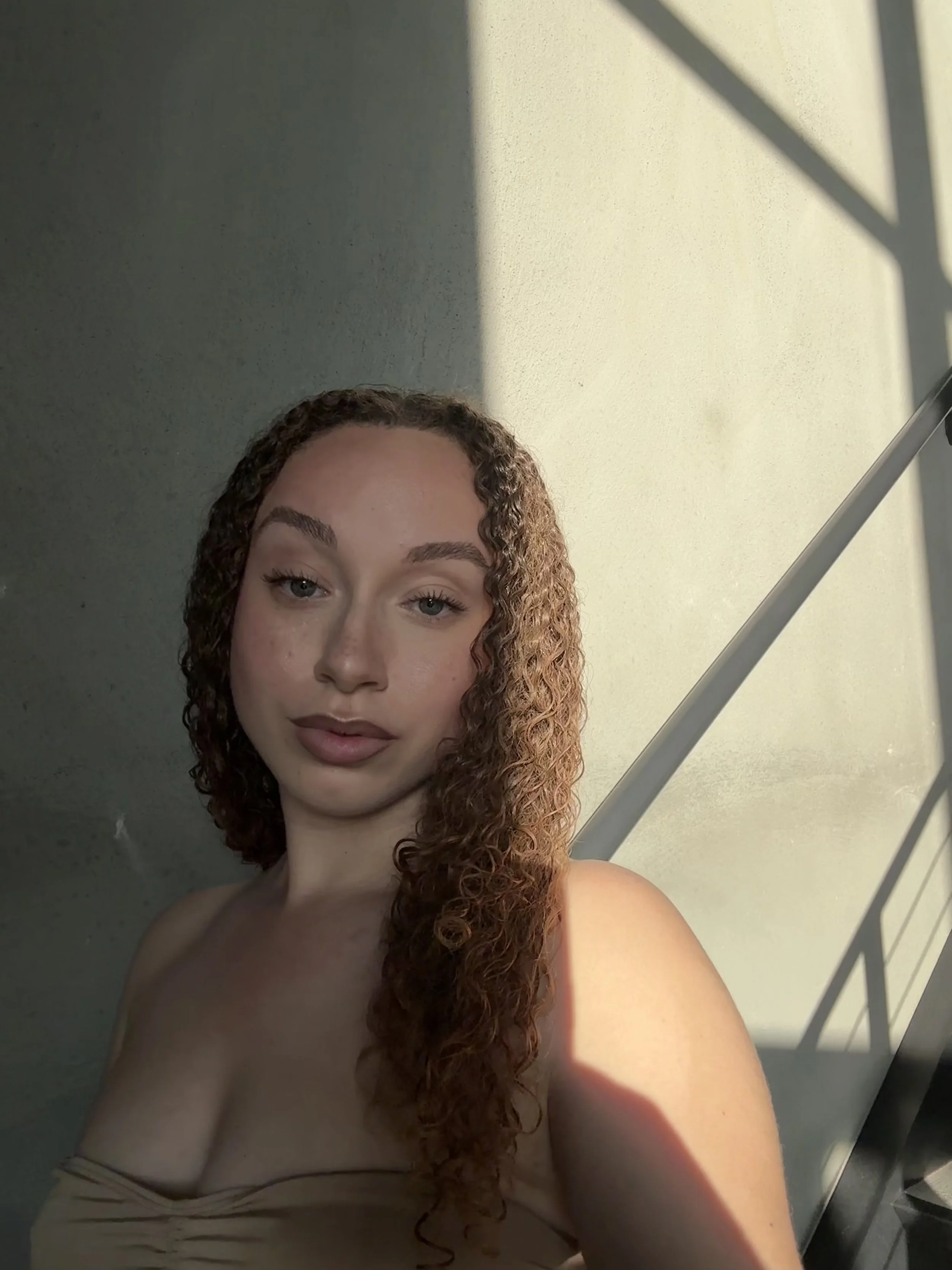 A young woman with curly red hair and light skin takes a selfie near a stairwell, with sunlight casting shadows on the wall behind her.