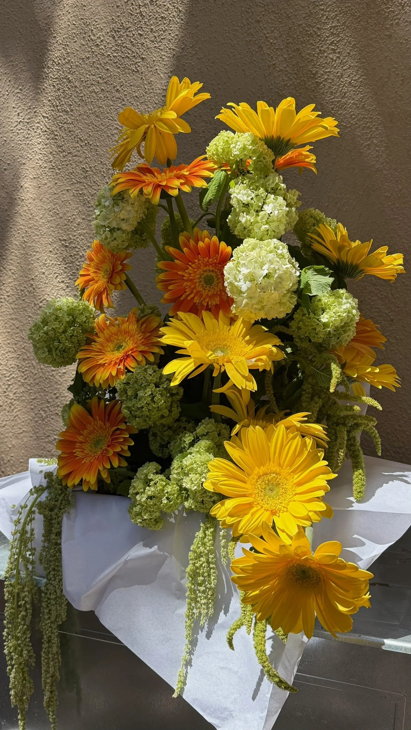A colorful flower arrangement in a white wrapping paper with yellow, orange, and white flowers, placed on a surface against a textured wall background.