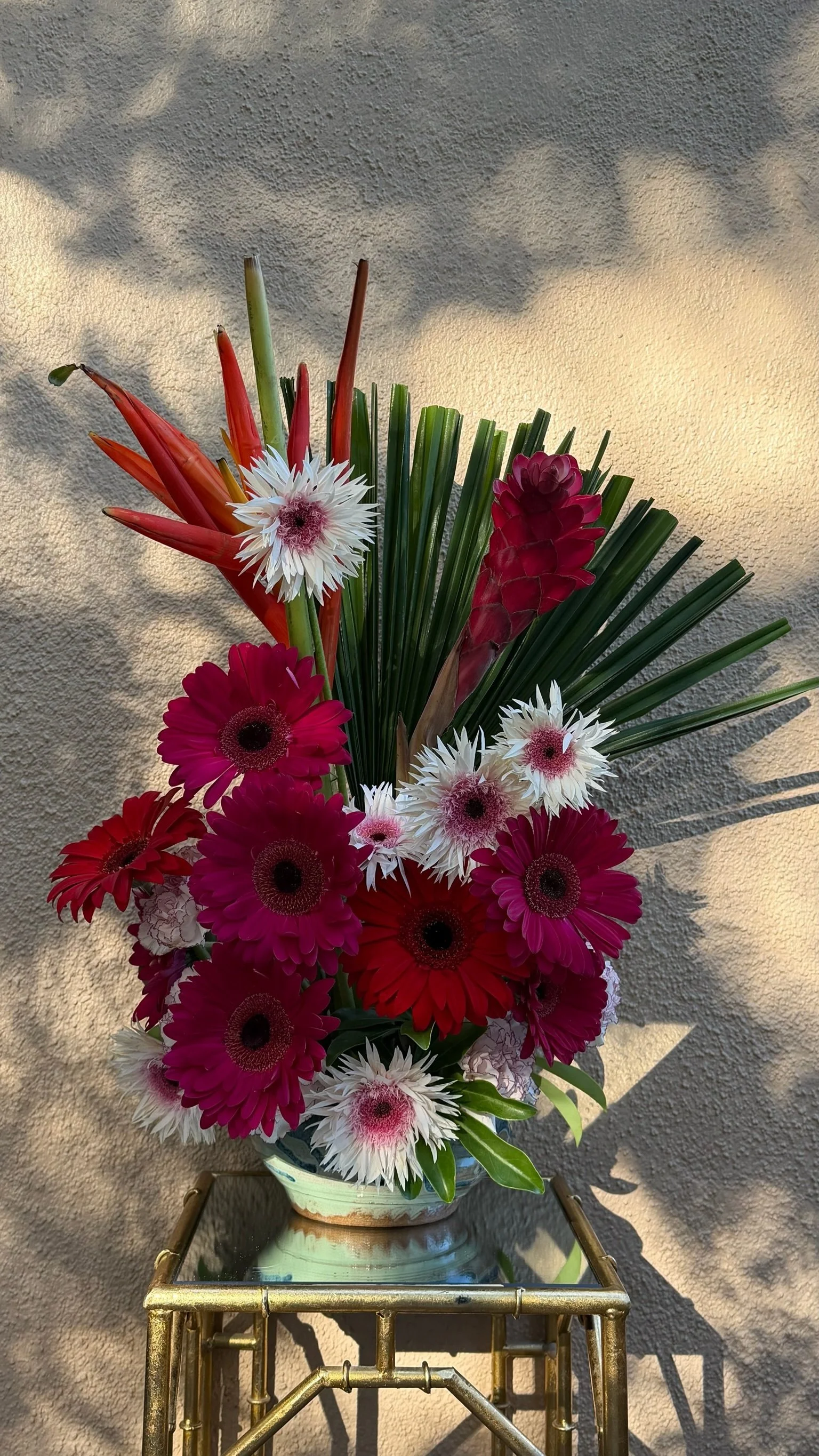 A colorful bouquet of flowers in a ceramic vase on a metallic gold table against a textured wall, illuminated by sunlight creating shadows.