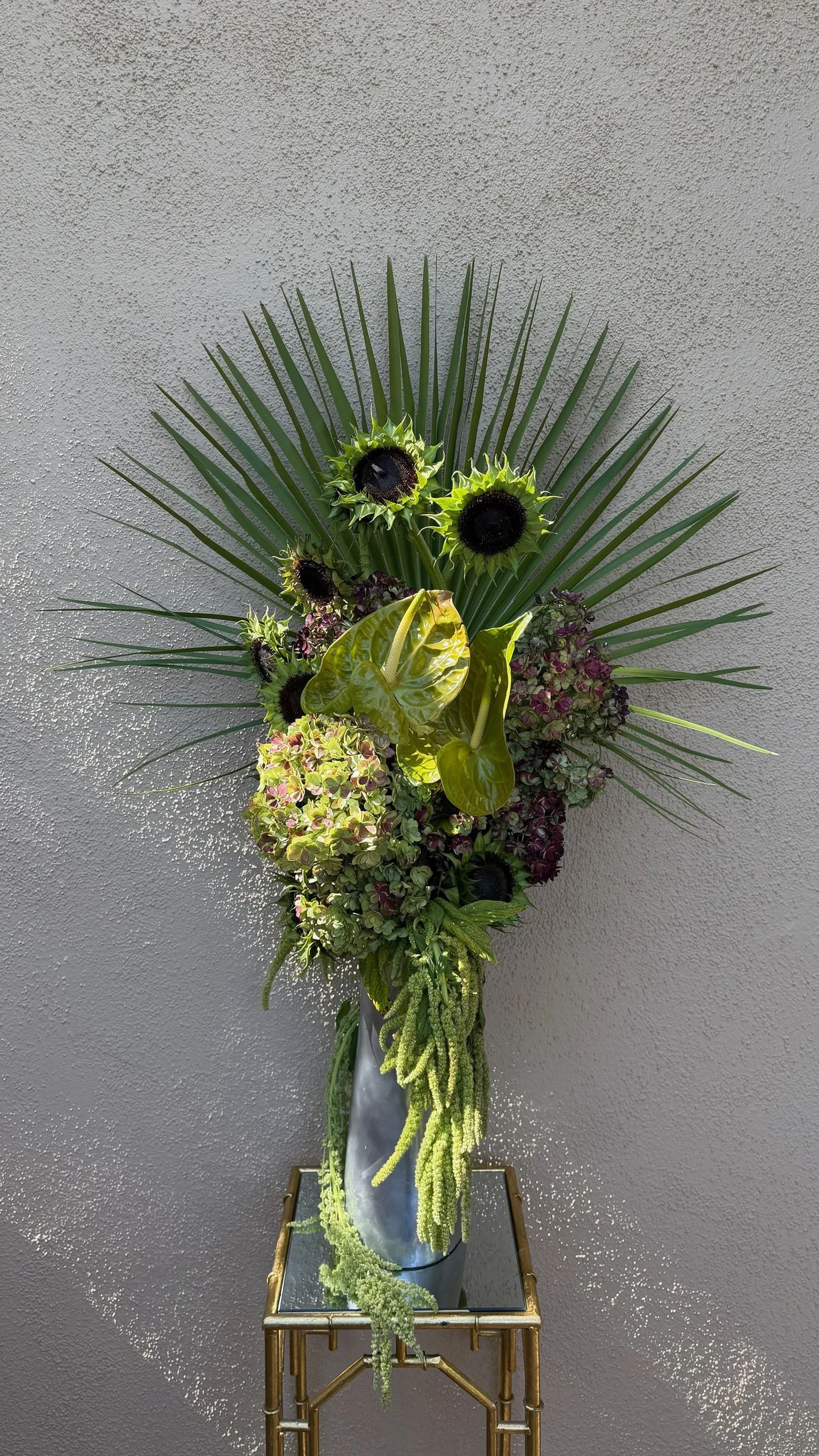 A tall floral arrangement in a silver vase on a gold metal stand against a textured gray wall. The arrangement includes green palm leaves, sunflowers, anthuriums, hydrangeas, and various green foliage.