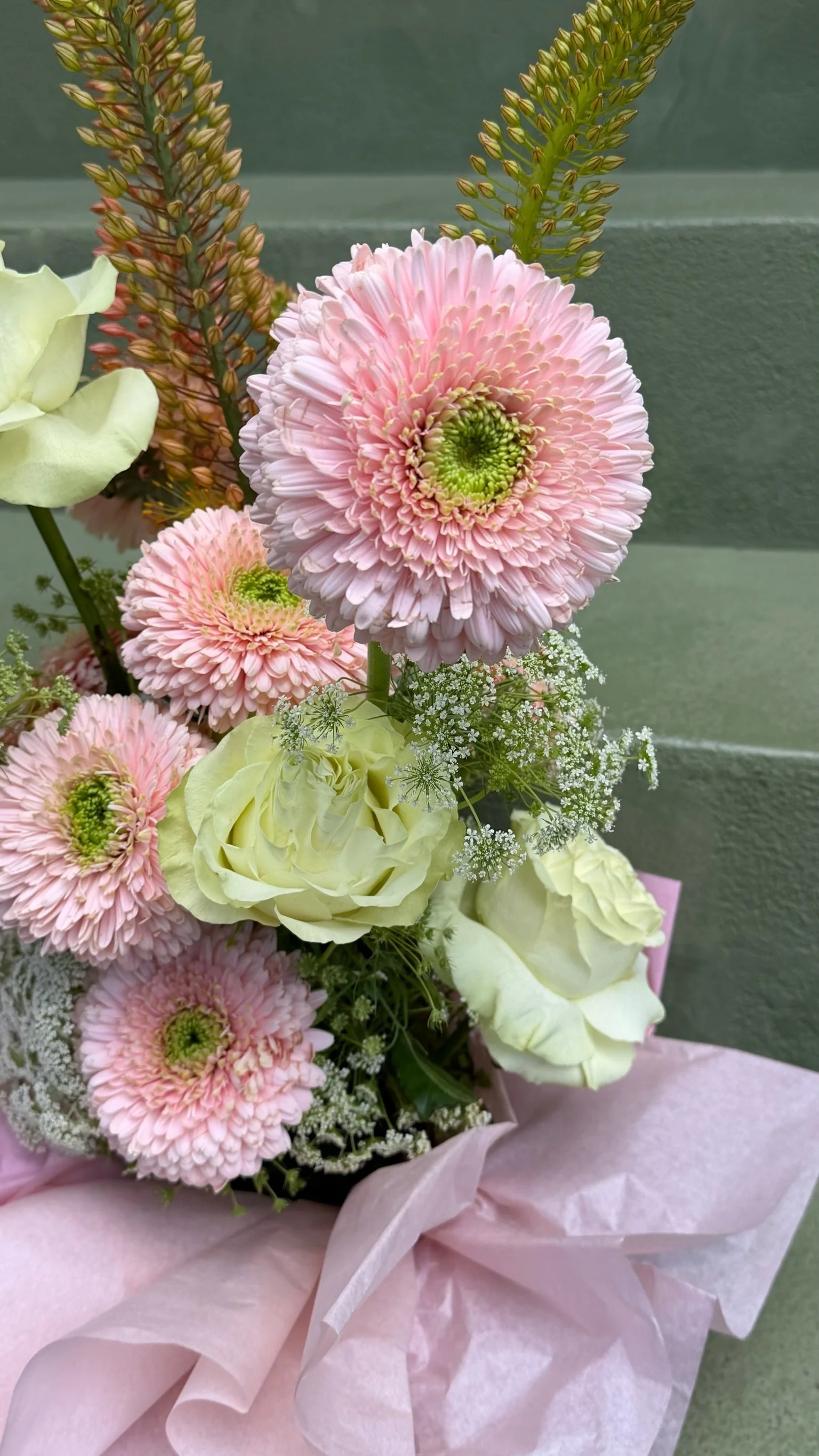Arrangement of pink daisies, white roses, and small white filler flowers wrapped in pink tissue paper.