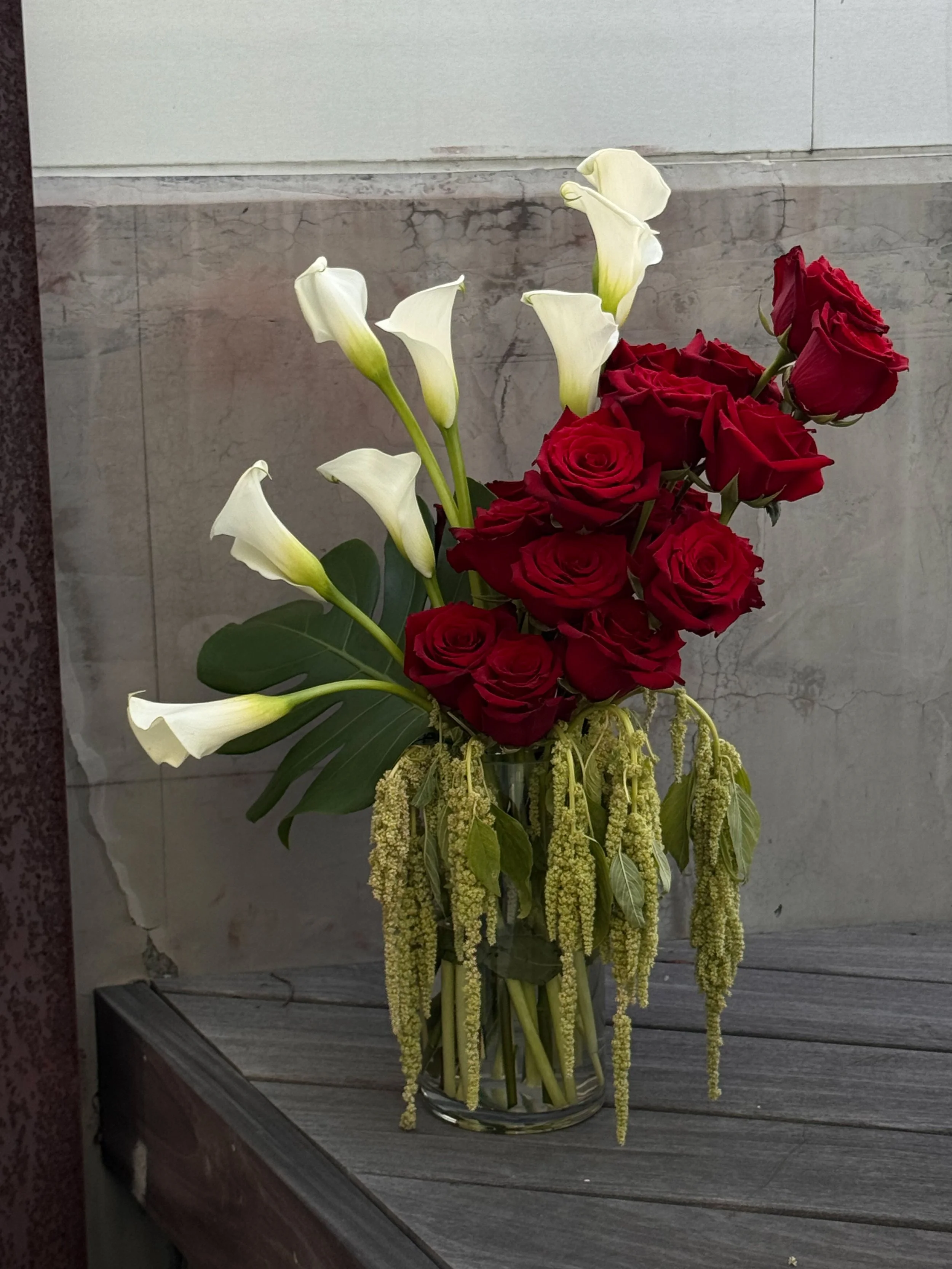A bouquet of white calla lilies and red roses arranged in a clear glass vase on a wooden surface with a marble wall background.