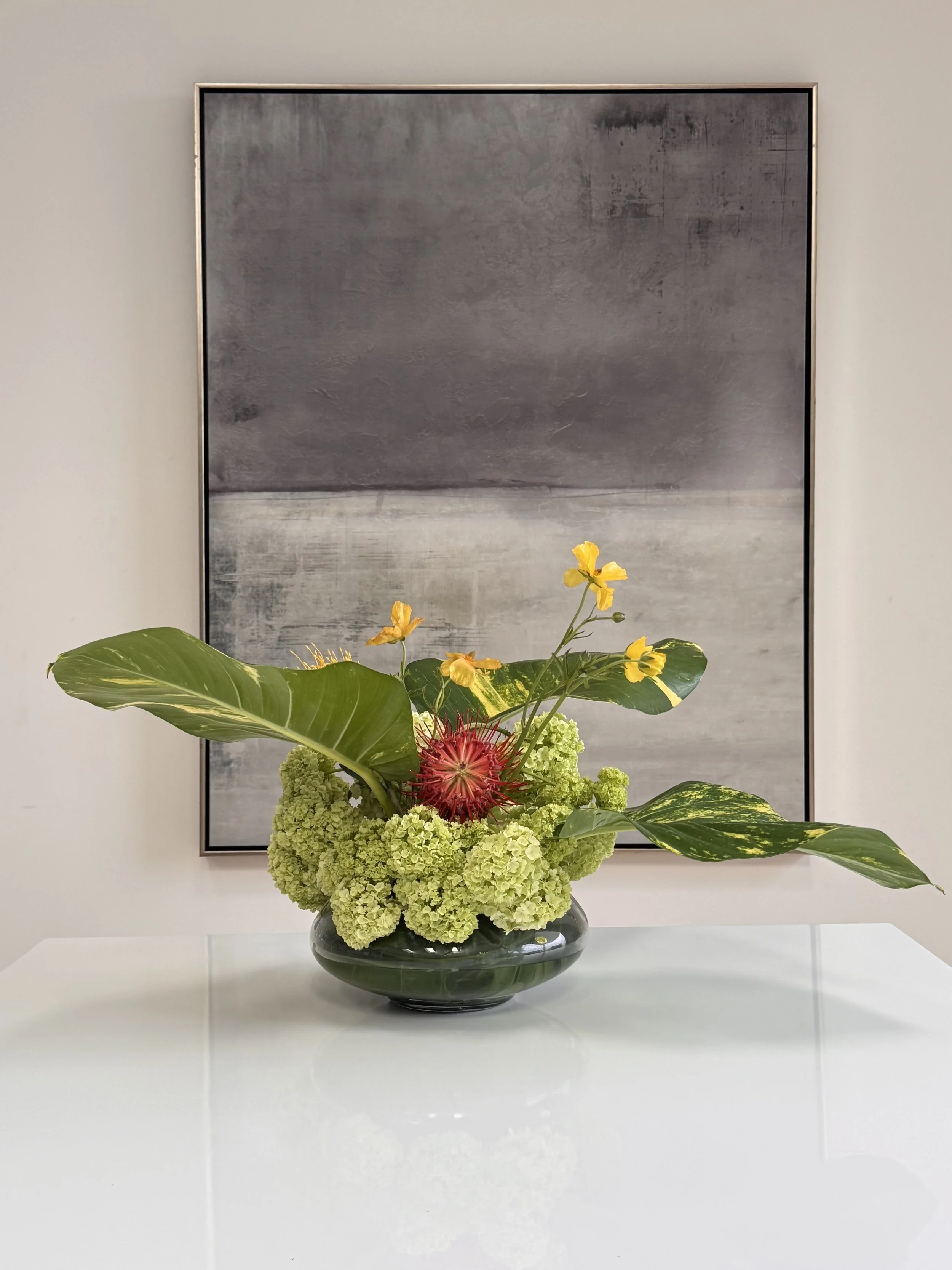 A black vase filled with green hydrangeas, yellow orchids, red protea, and green leaves, placed on a white table in front of a large abstract painting.