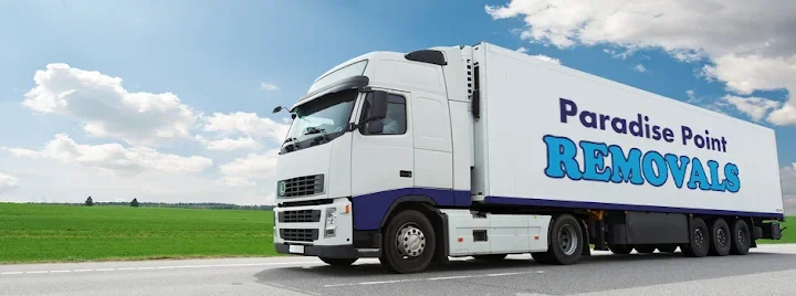 A large white moving truck with the words 'Paradise Point Removals' on the side driving on a highway with a green field and blue sky in the background.