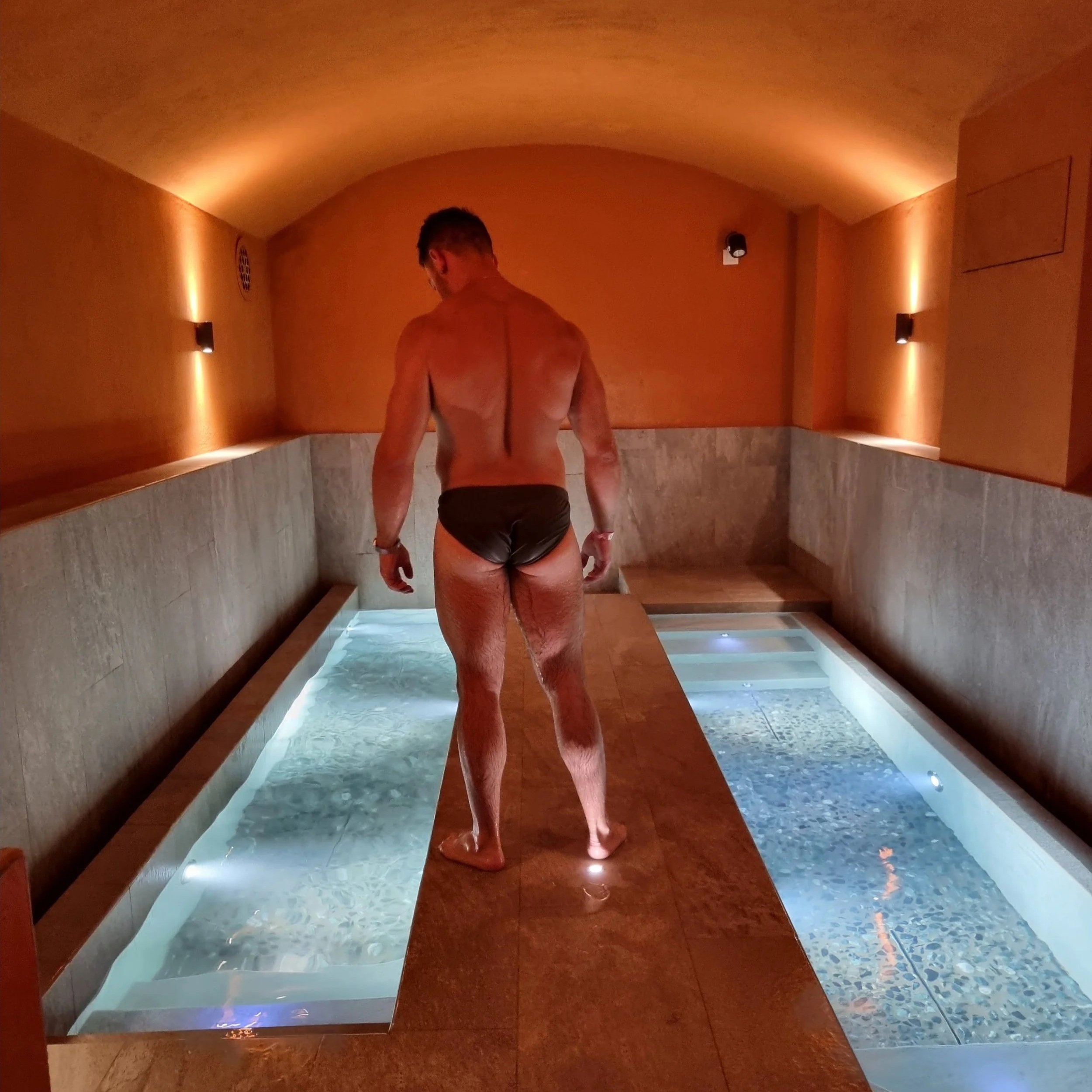 Uomo nudo che si trova in una stanza con pareti color terra e luci soffuse, vicino a una vasca o piscina a bordo raso.