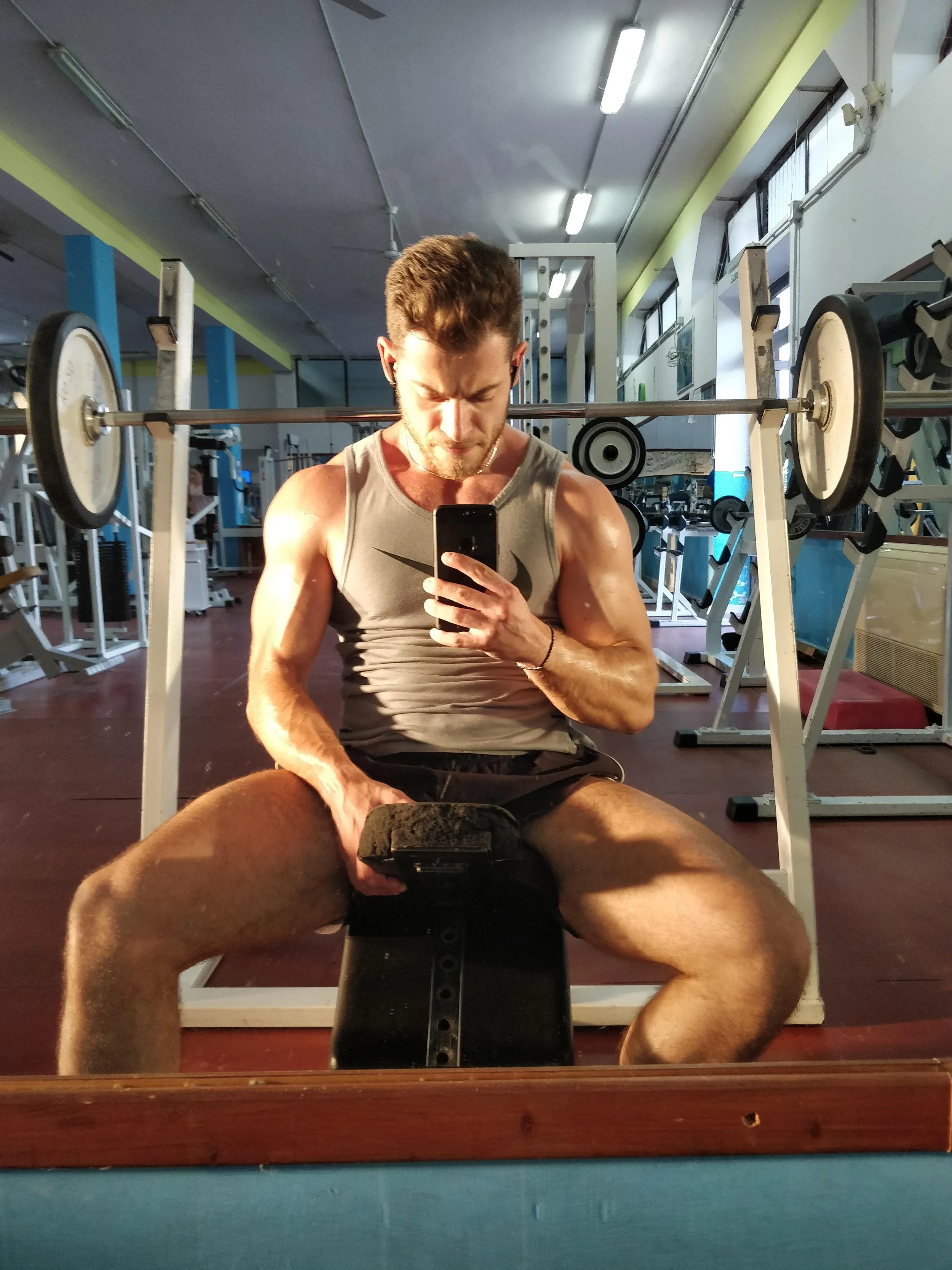 Un uomo muscoloso seduto in palestra, con un bilanciere davanti, che si scatta un selfie allo specchio. L'ambiente è ben illuminato e ci sono altri attrezzi da palestra sullo sfondo.