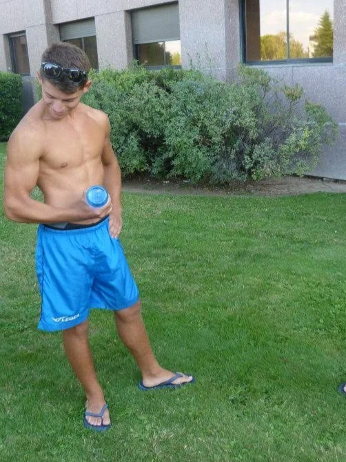 Giovane uomo con pantaloncini blu e infradito, con occhiali da sole sulla testa, guarda una bottiglia blu mentre si trova sul prato verde all'aperto.