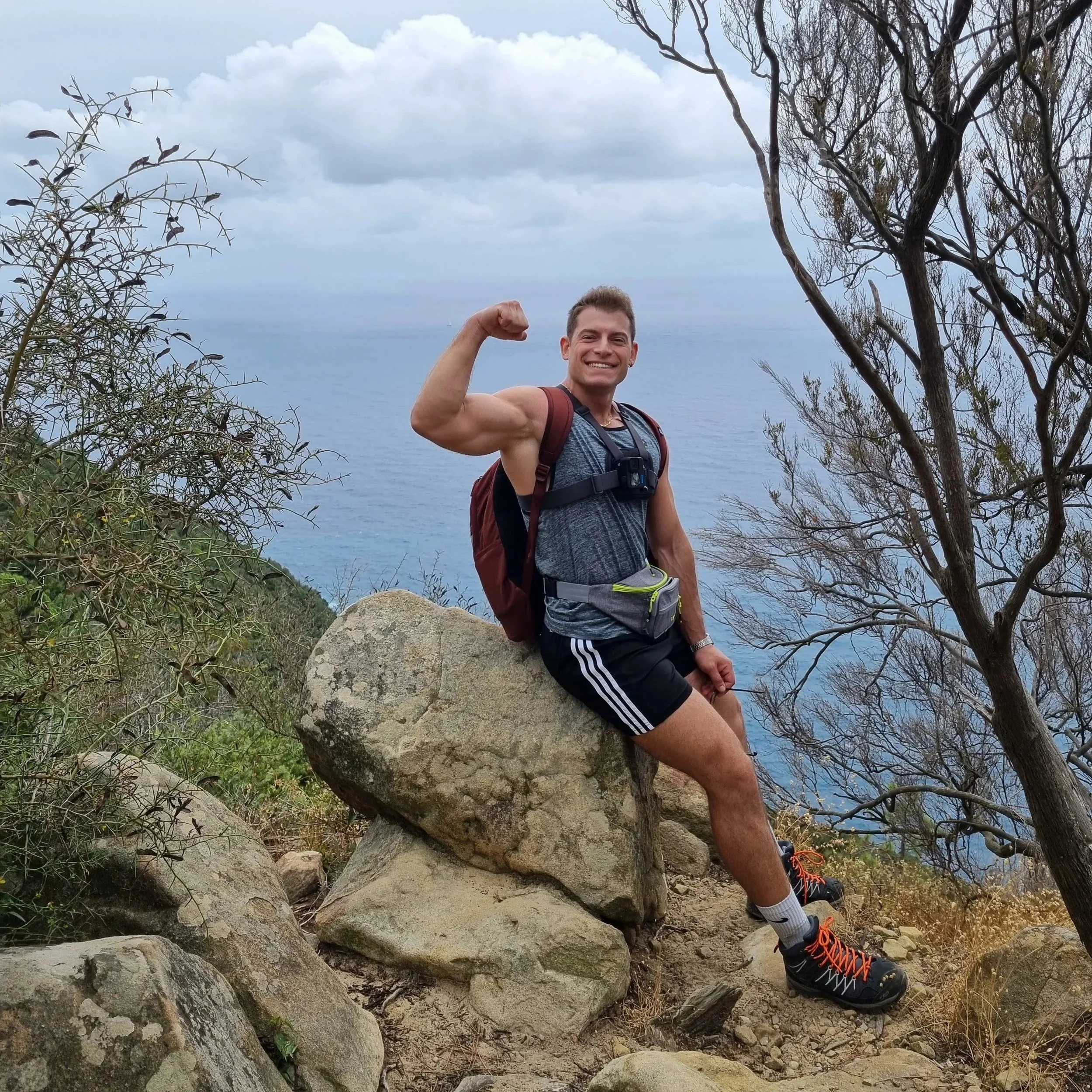 Un uomo sorridente con una maglietta grigia, pantaloncini neri con strisce biane, scarpe da trekking nere con dettagli arancioni, e uno zaino rosso in spalla, si trova in una zona rocciosa con alberi spogli sullo sfondo e il mare visibile in lontanan