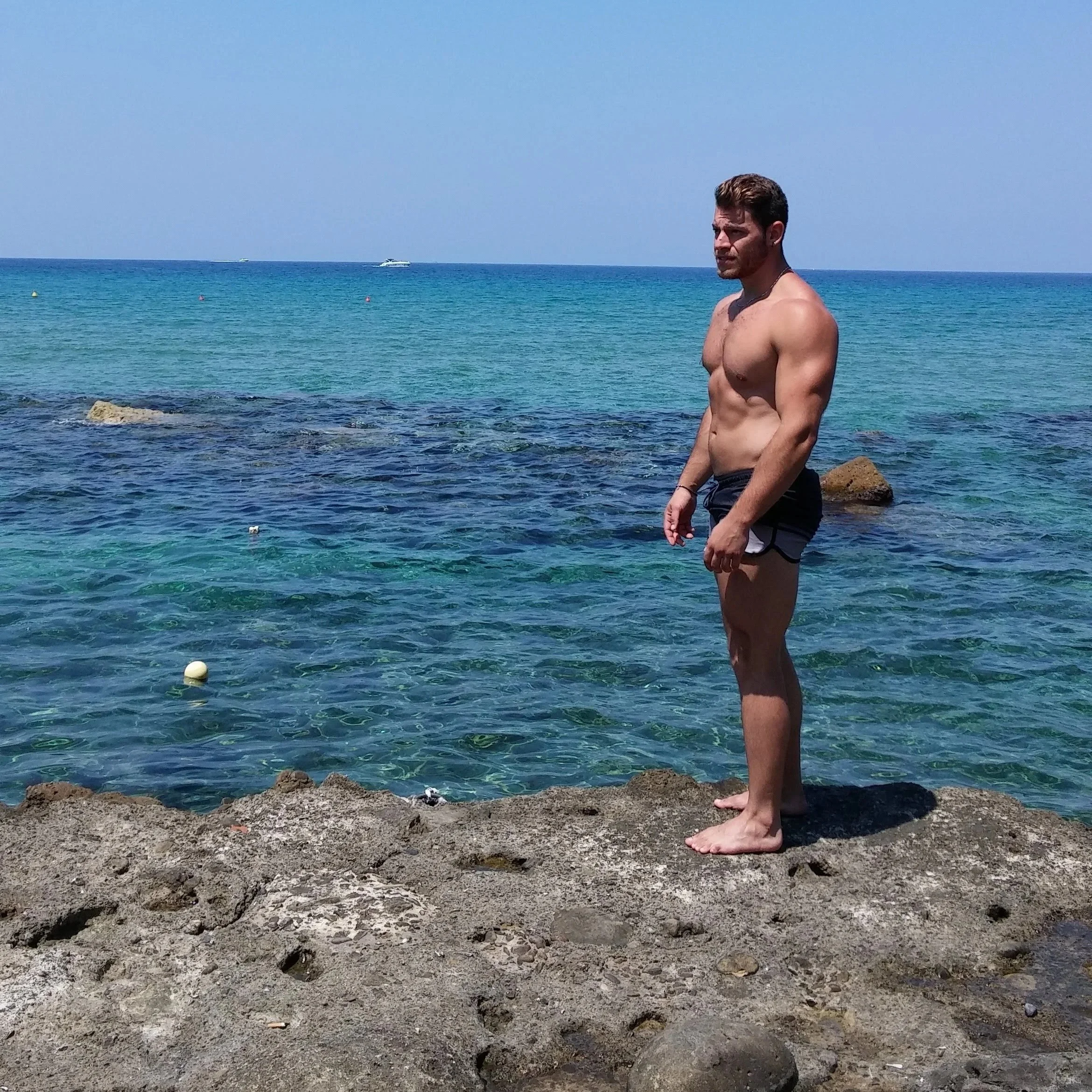 Uomo a torno nudo che si trova sulla riva del mare con acqua cristallina azzurra, con poche barche visibili in lontananza e rocce sulla spiaggia.