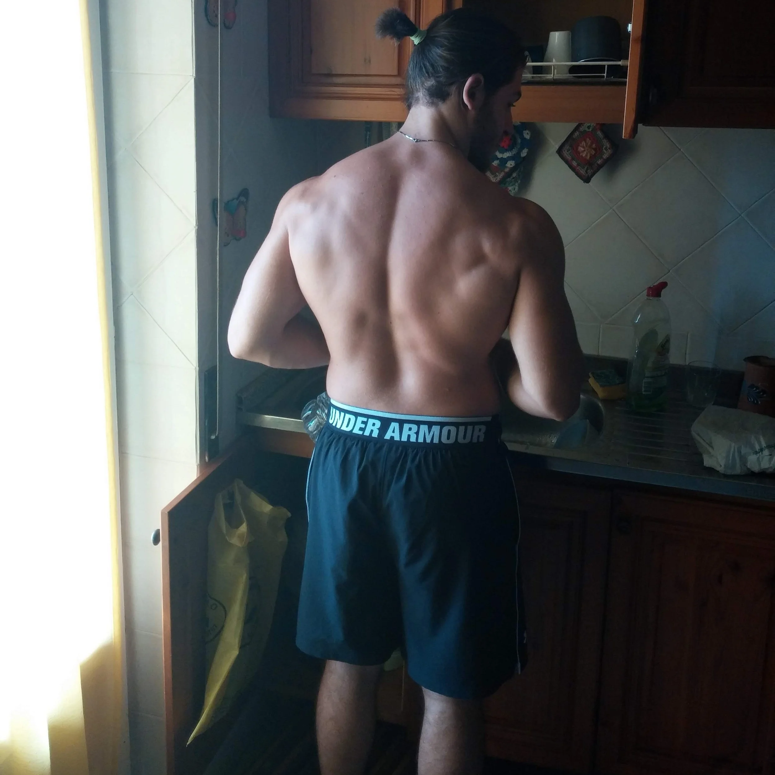 Un uomo senza maglietta, con i capelli raccolti in una coda, si trova in piedi, con le spalle e la schiena visibili, nella stanza con mobili in legno, un tappeto a motivi floreali e un pavimento in piastrelle. C'è una brocca di vetro sul tavolino sulla destra.