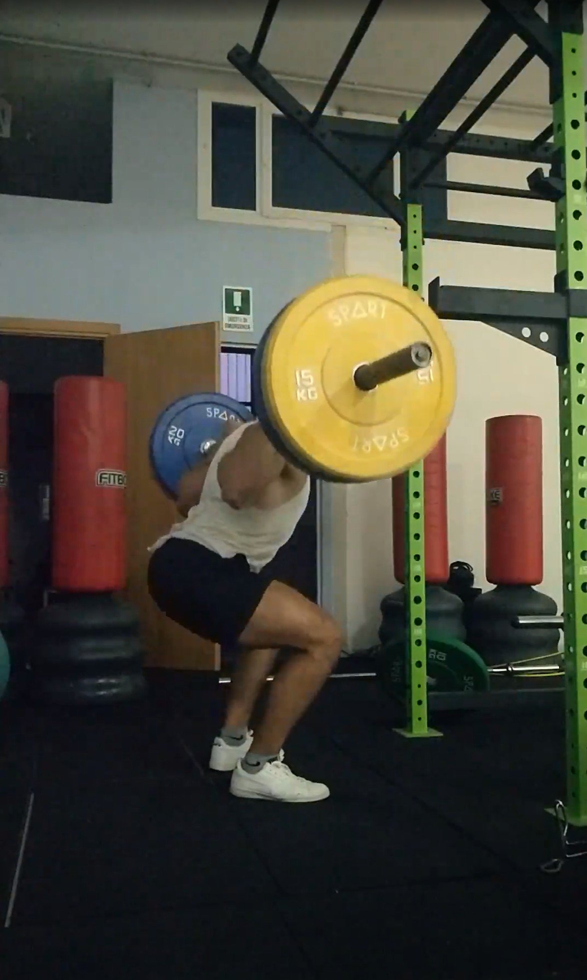 Una persona sta facendo squat sollevando un bilanciere con pesi gialli e blu in una palestra, circondata da sacchi da boxe e attrezzature da allenamento.
