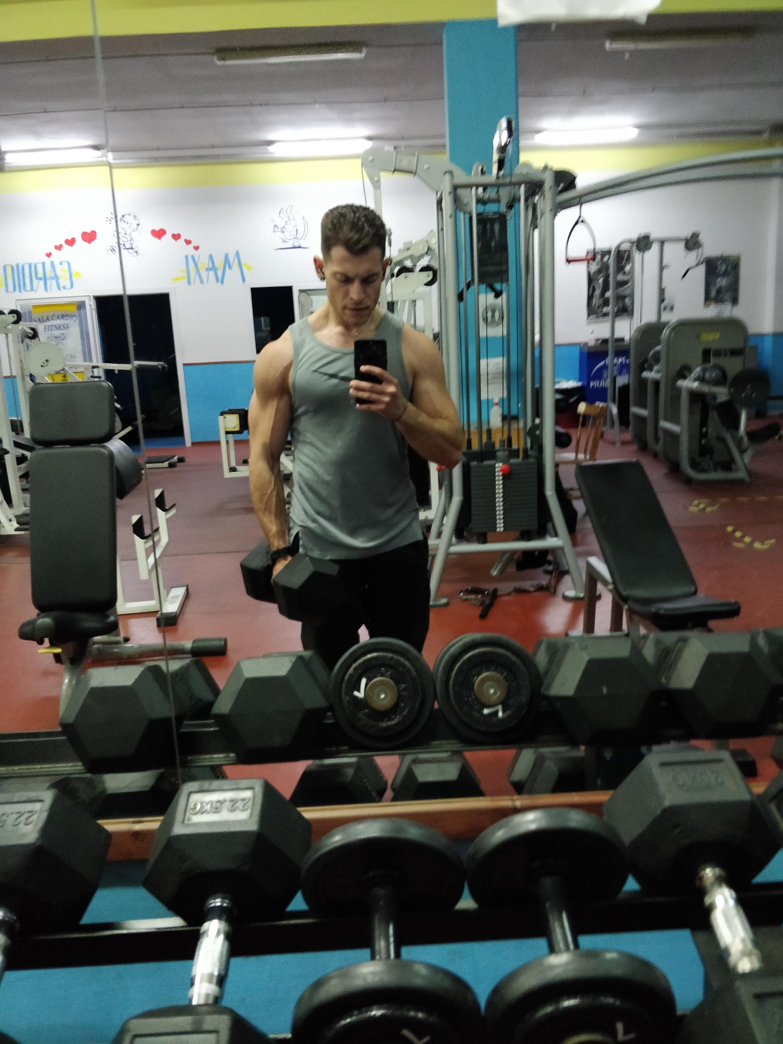 Ragazzo che si allena in palestra, si guarda allo specchio mentre prende un manubrio, con attrezzature da palestra intorno e un muro decorato con disegni e scritte.