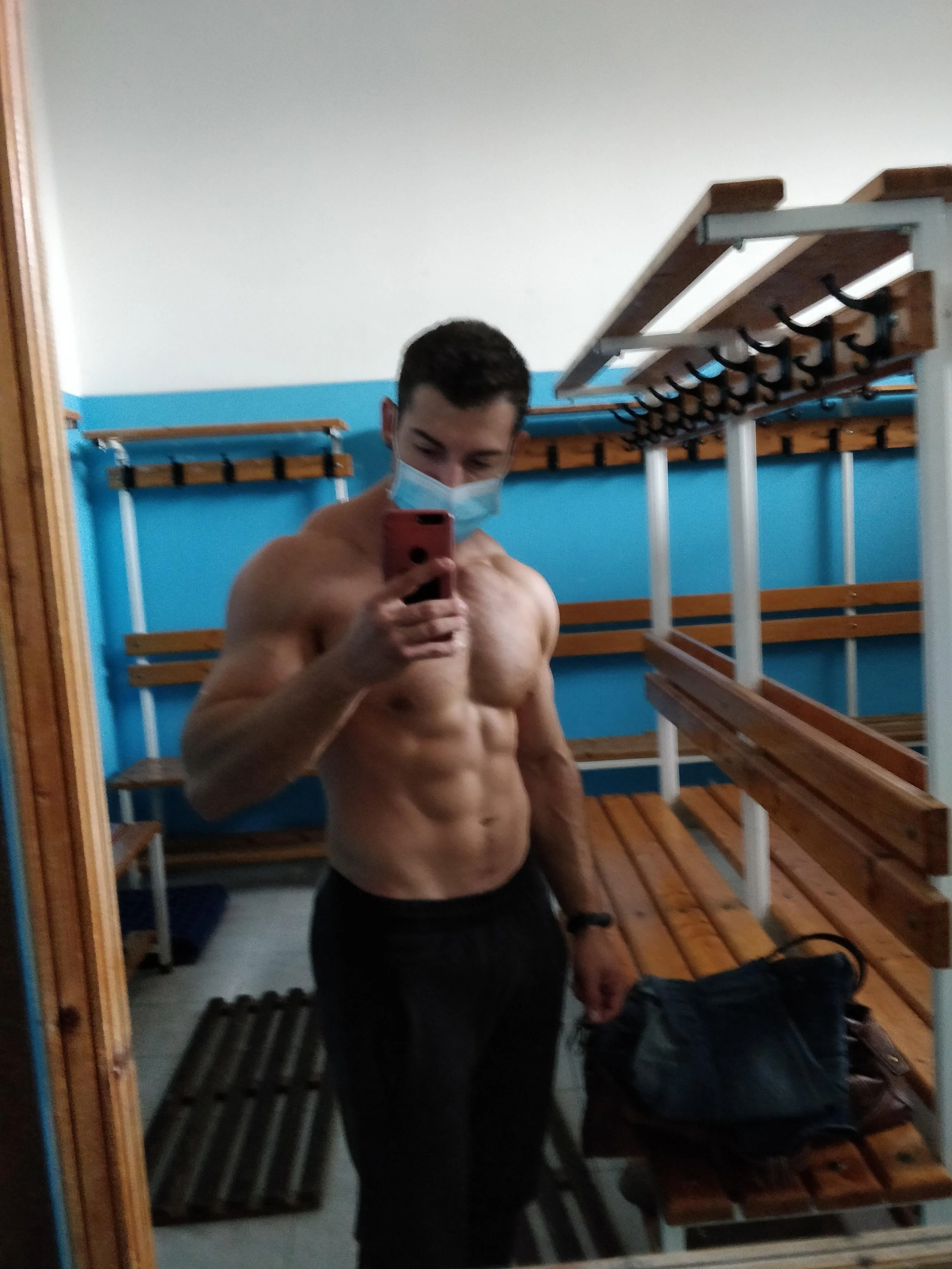 Un giovane uomo senza maglietta, indossa una mascherina e si sta facendo un selfie in uno spogliatoio, con armadietti di legno e panche sullo sfondo.