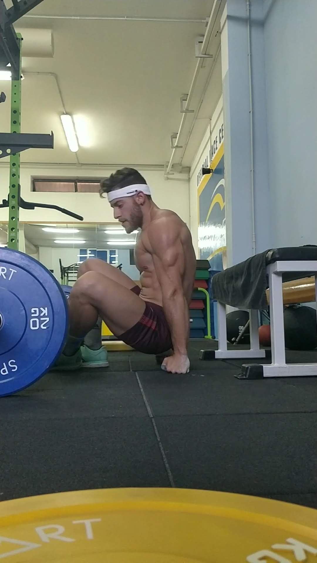 Uomo in palestra che si esercita in una posa di isometria muscolare, con muscoli evidenti e senza maglietta, indossa pantaloni corti color burgundy e una fascia bianca sulla testa.
