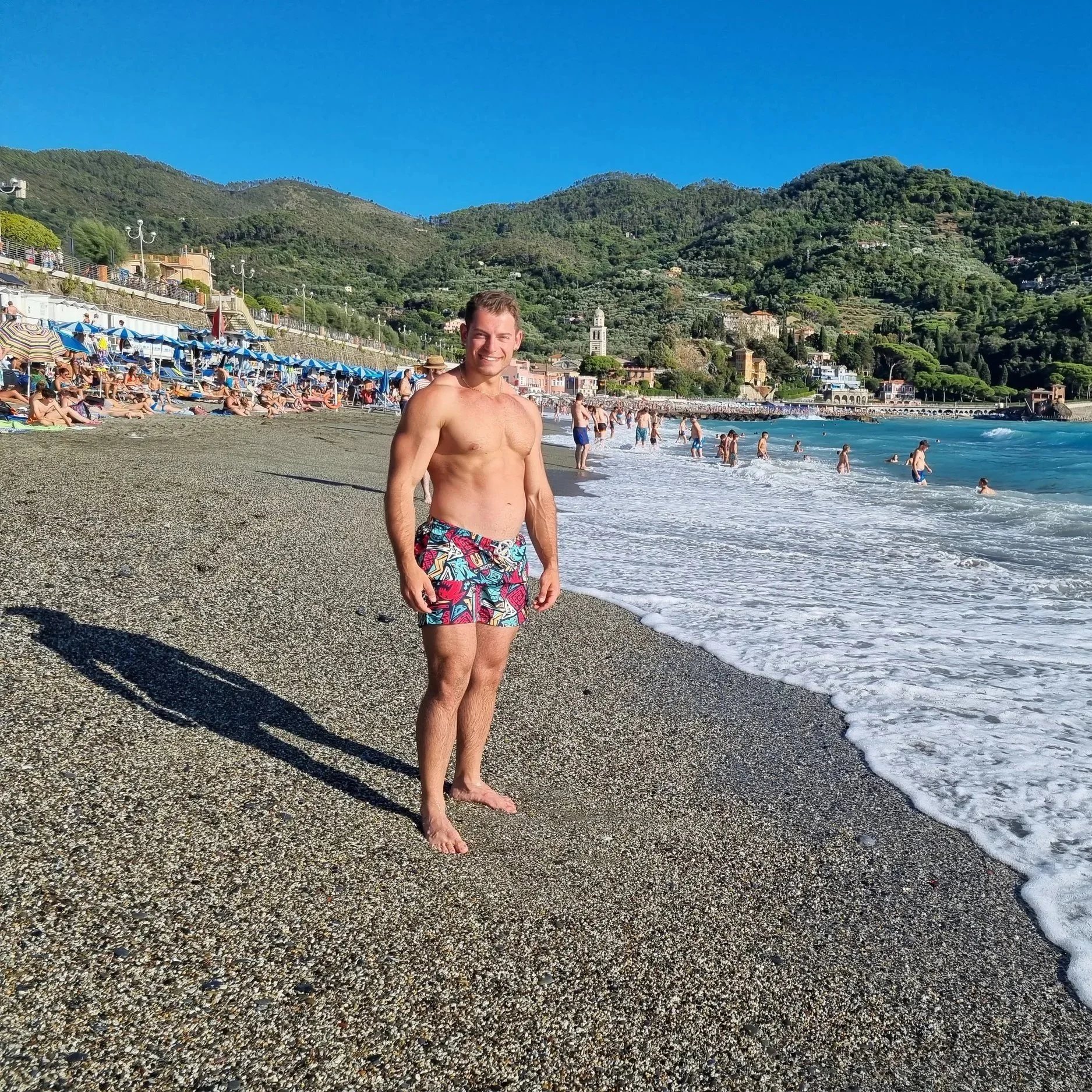 Uomo in costume da bagno con pantaloncini colorati sulla spiaggia, con oceano e montagne sullo sfondo, molte persone che si rilassano e nuotano in mare.
