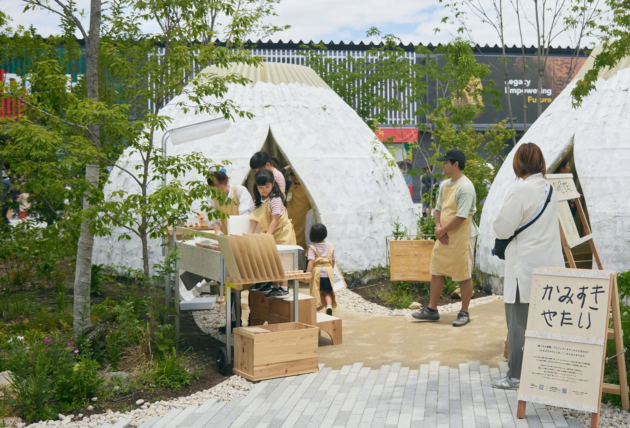屋外の展示会場で木製の作品を販売しているグループと購入者の人々。背景には緑の木やパネル看板があり、青空が見える。