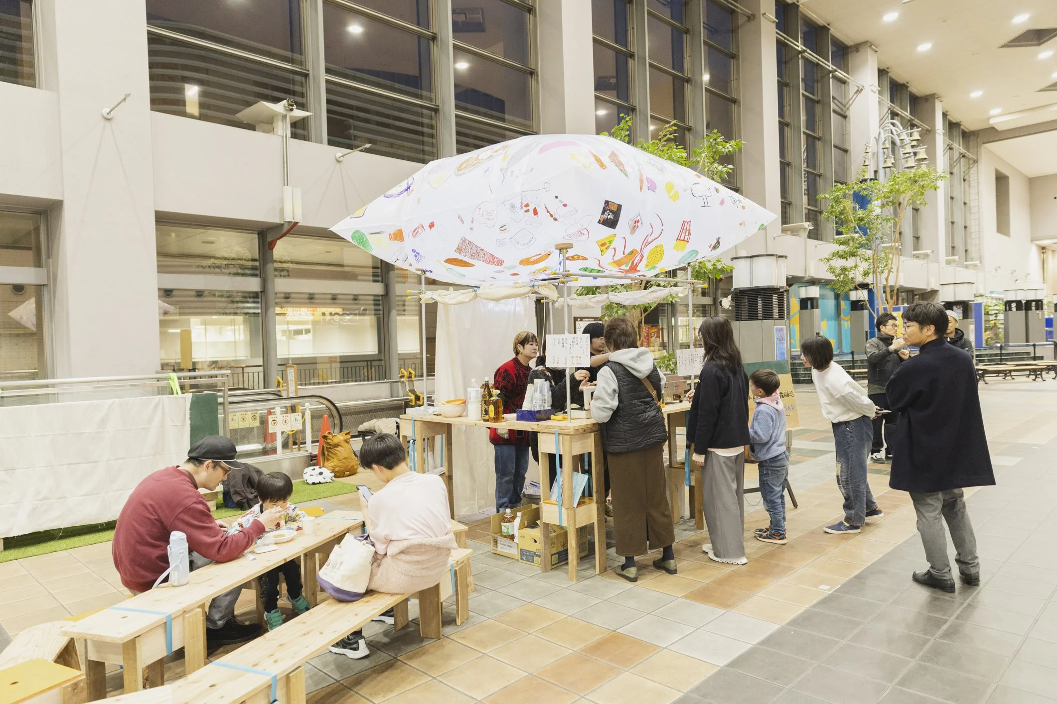屋内の広い会場で、多くの人々が屋台やブースに集まり、手作り品や飲み物を販売している。天井には大きなステンドグラスの模様のパラソルが吊るされている。