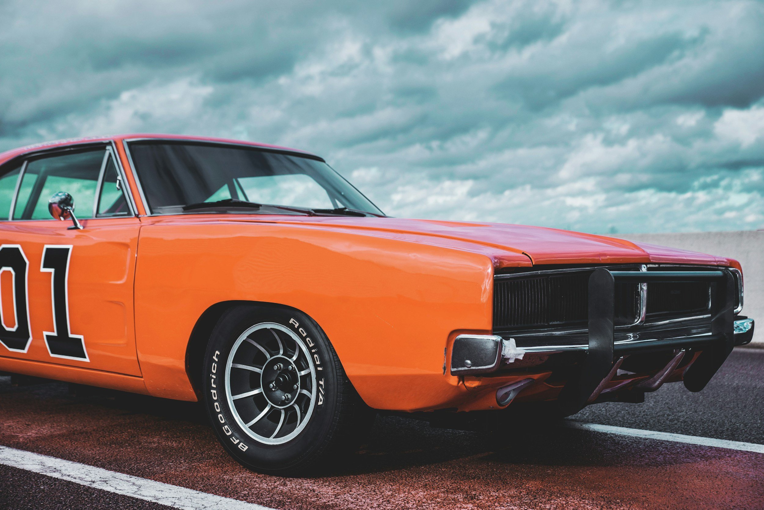 Orange vintage race car with black tires and a black front grill, parked on a blacktop surface under a cloudy sky.