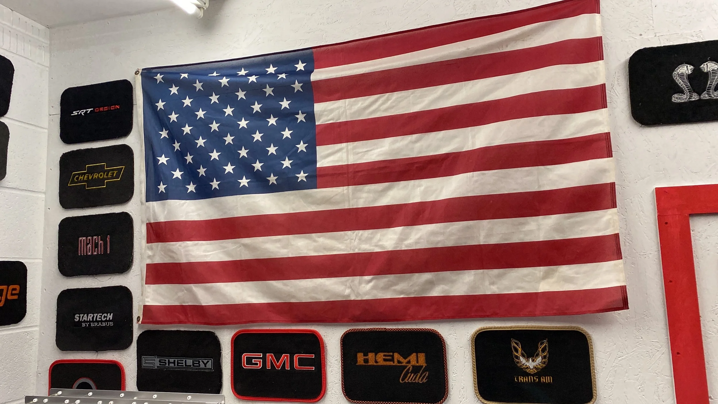 American flag hanging on a wall with automotive brand patches around it, including Chevrolet, GMC, Shelby, and Hemi.